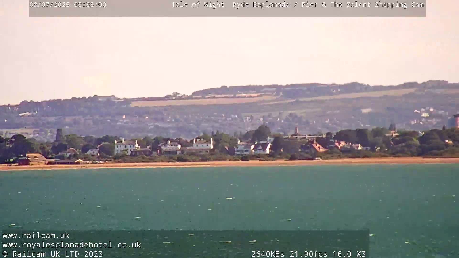 A calm body of teal water stretches towards a sandy beach and a line of buildings backed by distant hills under a hazy sky.