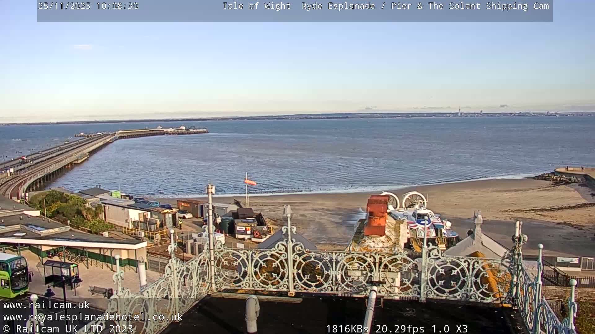 A sunny coastal scene features a long pier extending into calm blue water, a sandy beach with a hovercraft on the right, and various buildings including a train station in the foreground, all under a clear sky.