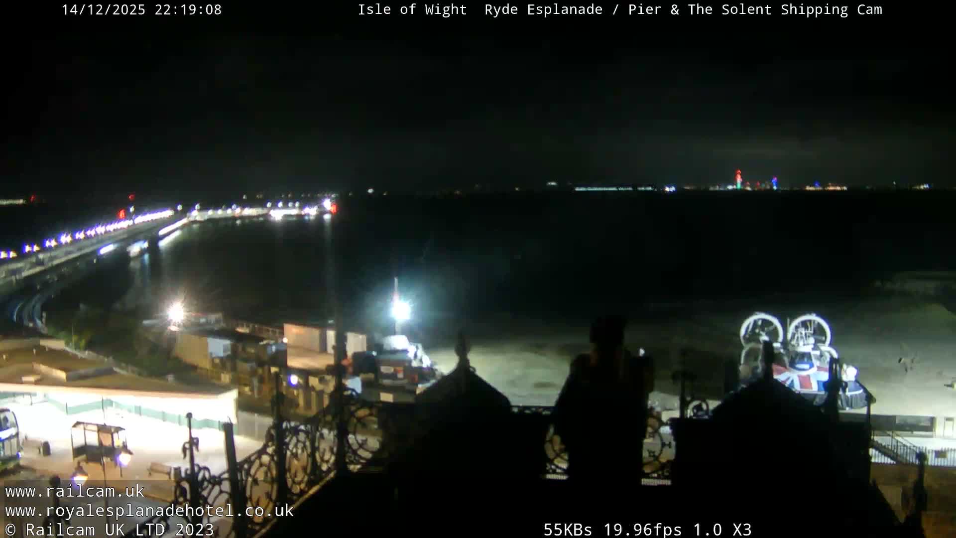 A sunny coastal scene features a long pier extending into calm blue water, a sandy beach with a hovercraft on the right, and various buildings including a train station in the foreground, all under a clear sky.