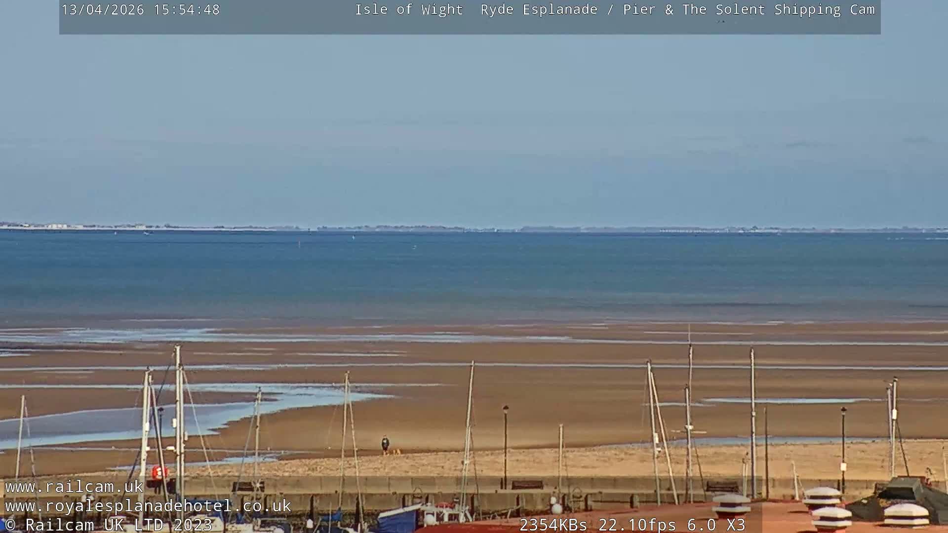 A sunny coastal scene features a long pier extending into calm blue water, a sandy beach with a hovercraft on the right, and various buildings including a train station in the foreground, all under a clear sky.