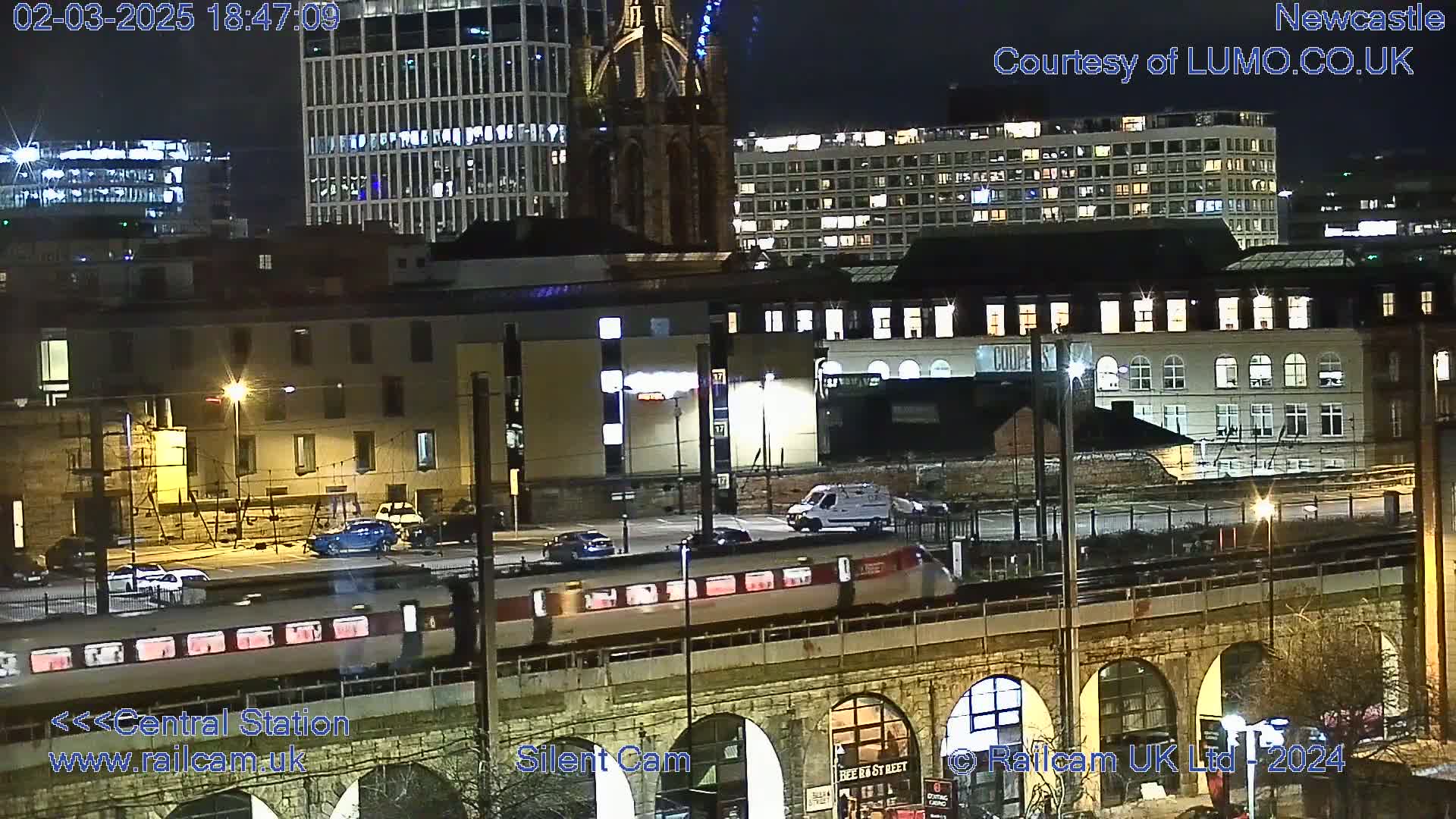 A nighttime view of a train passing over a stone arch bridge in a city, with buildings and streetlights illuminated.