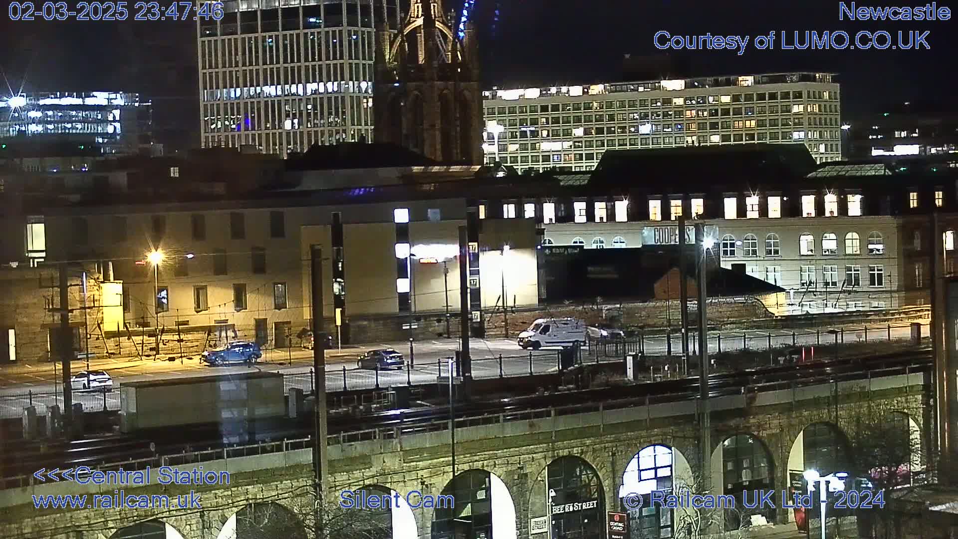 A nighttime view of a city street with several buildings, vehicles, and a stone arch bridge, under clear skies.