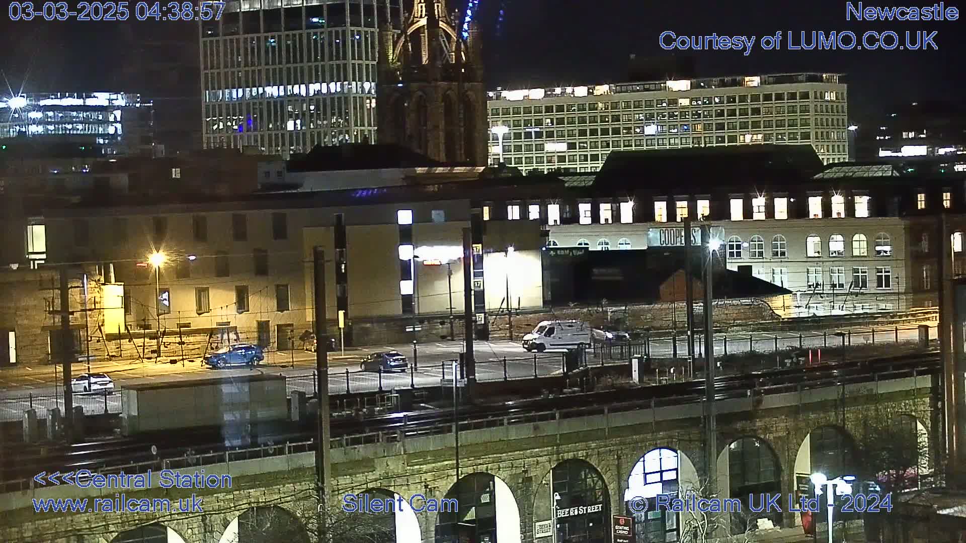 A nighttime view of a city's buildings and a parking lot, seen from a bridge over train tracks.