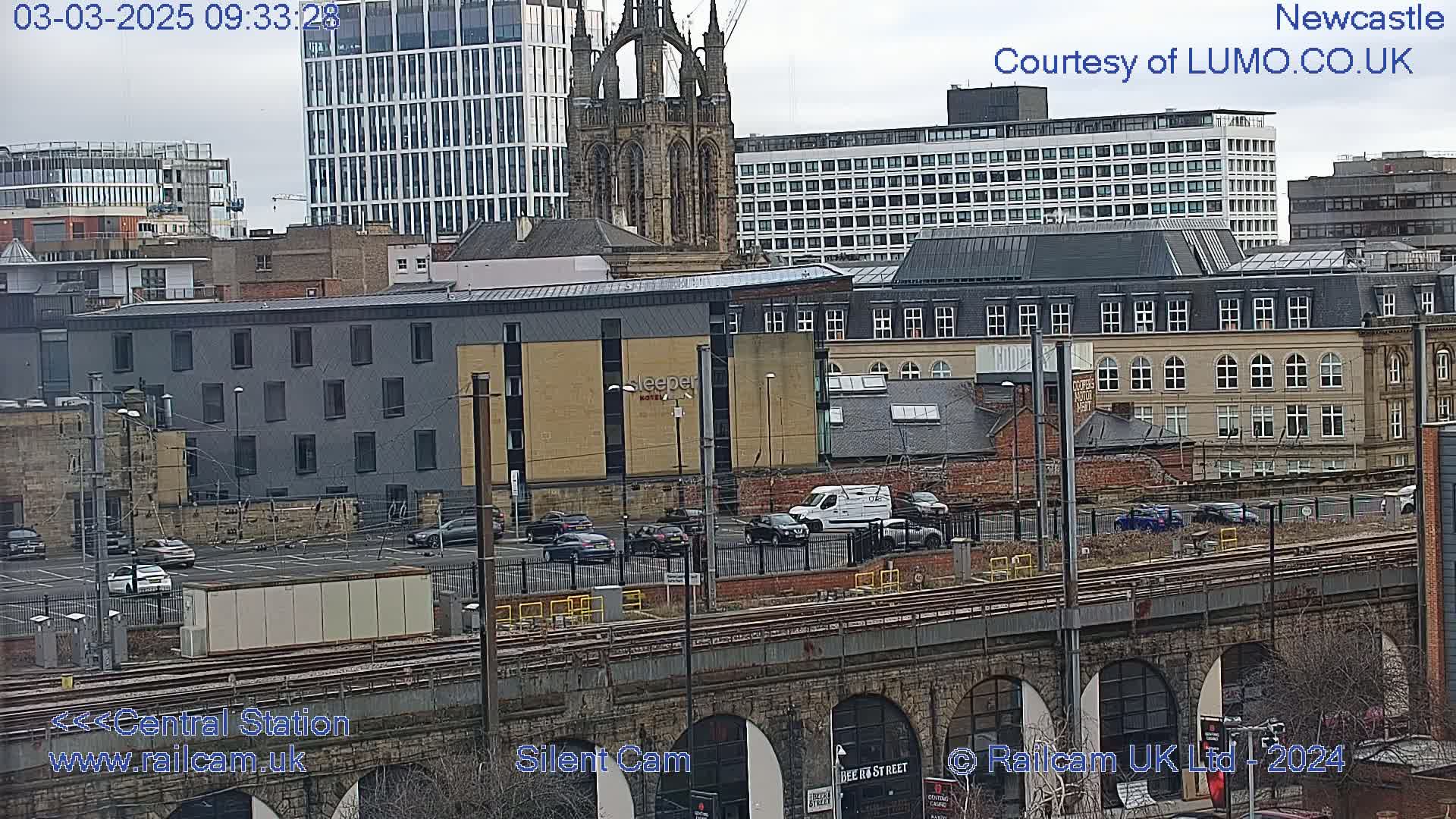 A view of a city on an overcast day, showing a railway viaduct, parking lots with several cars, and various buildings of different architectural styles, including a tall modern building and a gothic-style church.