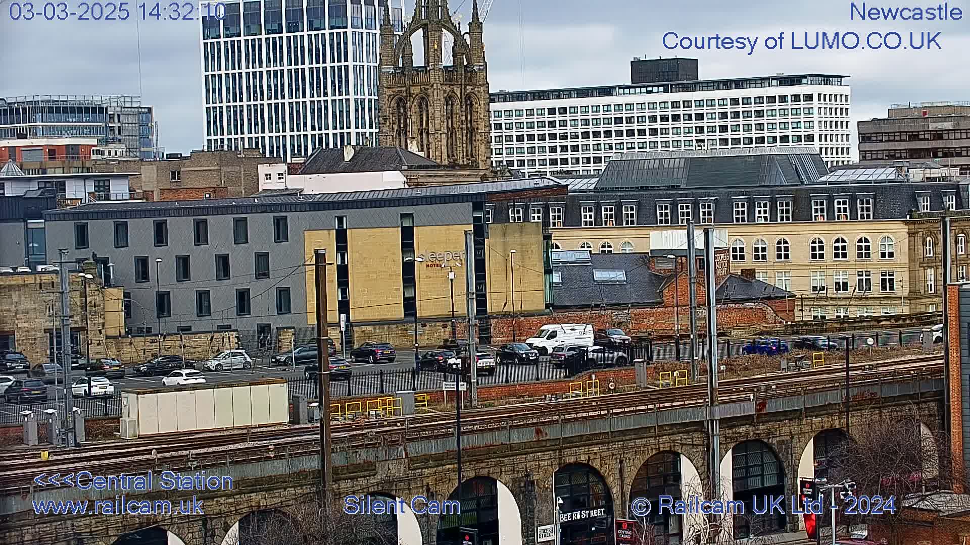 A city scene on an overcast day, viewed from above a railway viaduct, shows several buildings including a modern hotel, older brick structures, and a tall modern glass building, with cars parked in a lot below.
