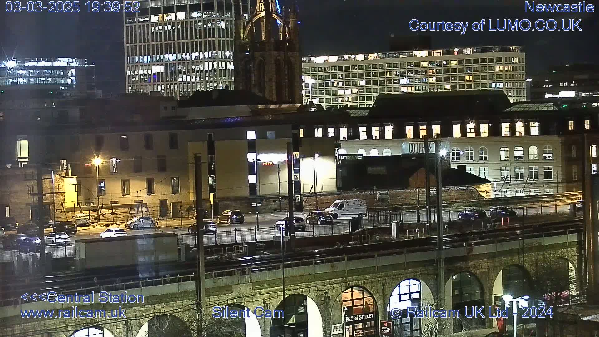 A nighttime view of a city's buildings and a parking lot near a train station under a clear sky.