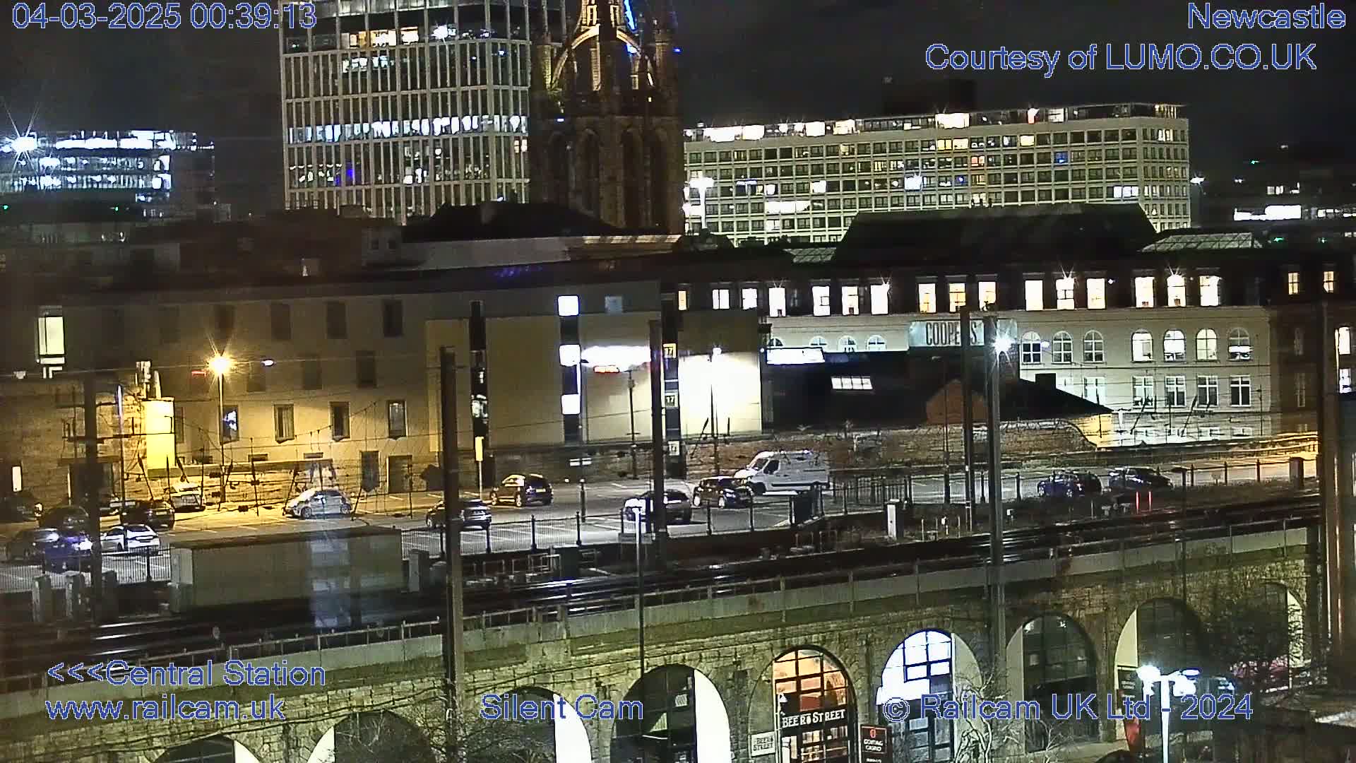 A nighttime view of a city's central station area, showing several buildings, parked cars, and a train track over a stone archway under a clear sky.