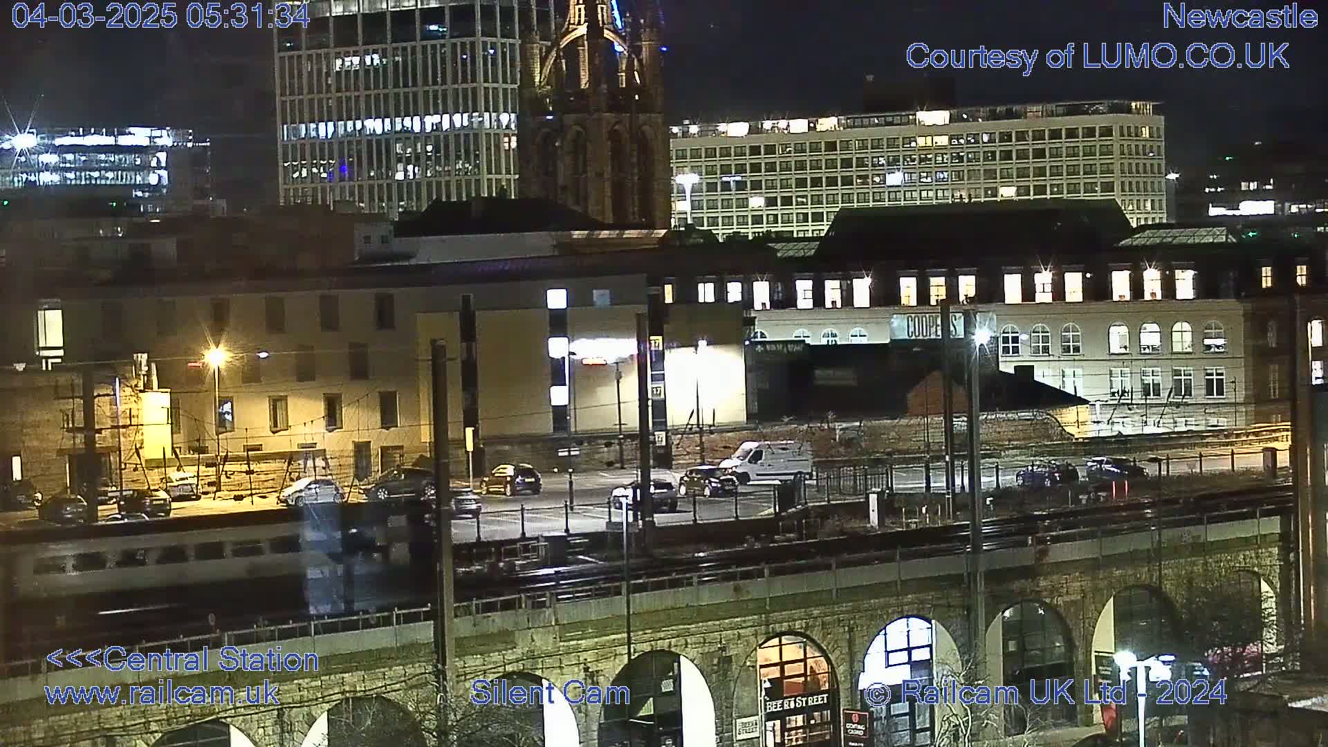 A nighttime view of a city train station and surrounding buildings, including a train on tracks and vehicles in a parking lot.