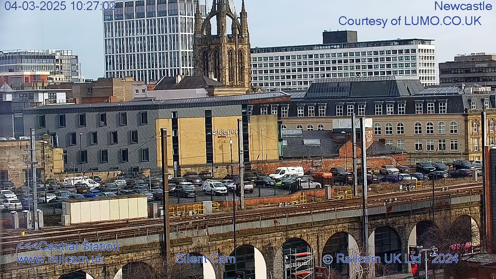 A sunny view of a city's cityscape, including several buildings, a parking lot full of cars, and train tracks on a stone viaduct.