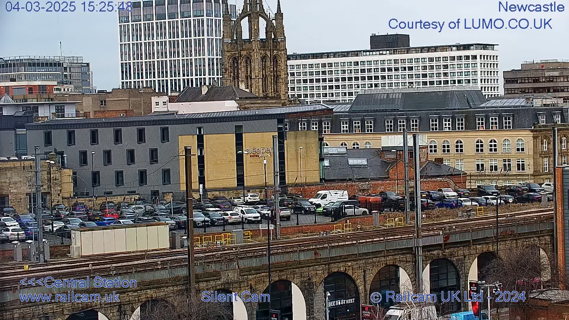 A city scene with overcast skies, showing a large parking lot full of cars next to a railway viaduct, with numerous buildings and a church spire in the background.