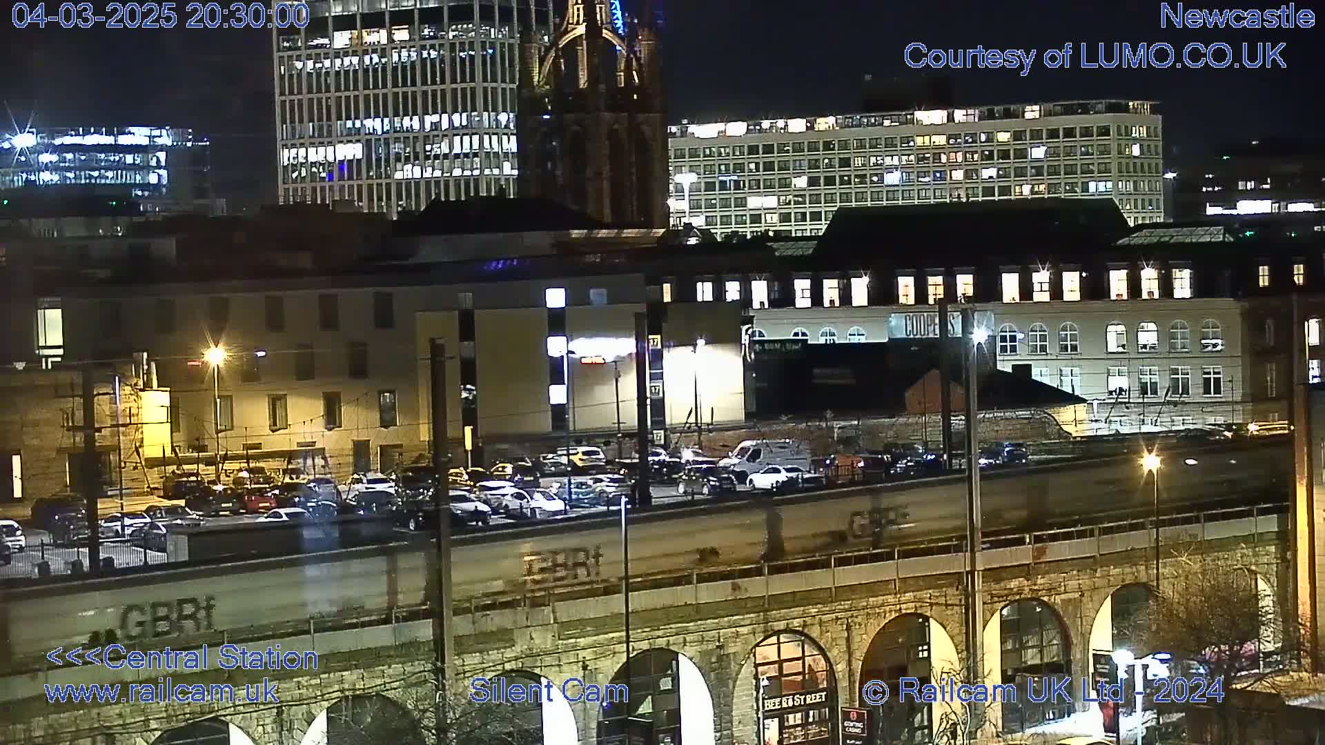 A nighttime view of a city, showing several buildings illuminated against a dark sky, with a train passing over a stone arched bridge below.