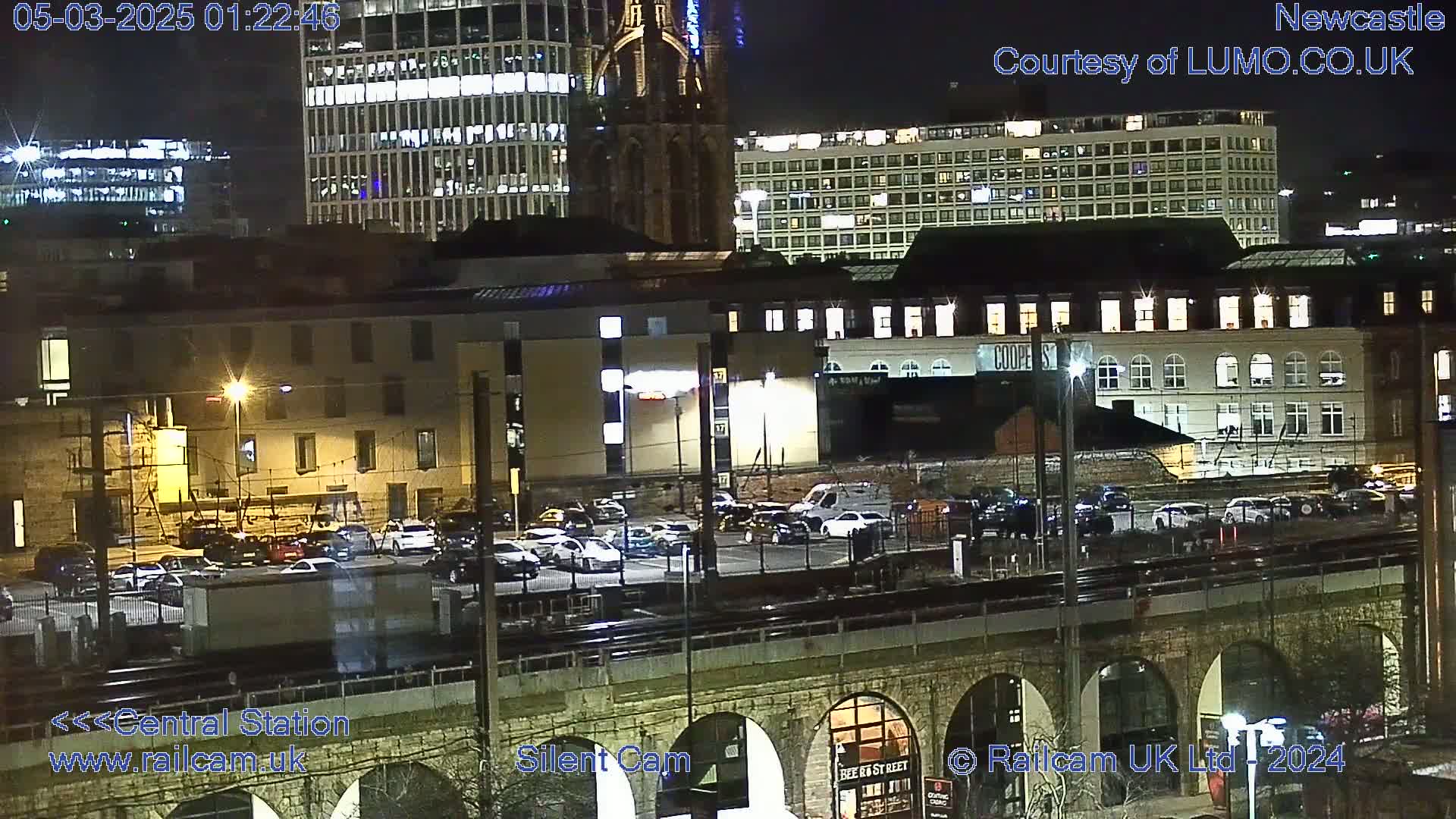 A nighttime view of a city train station and surrounding buildings, with cars parked in a lot below.