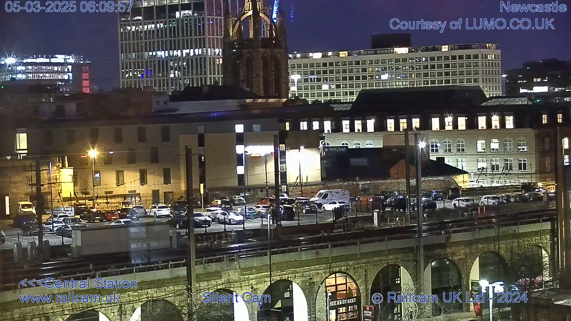 A nighttime view of a city’s railway station and surrounding buildings, including a parking lot, under a dark sky.
