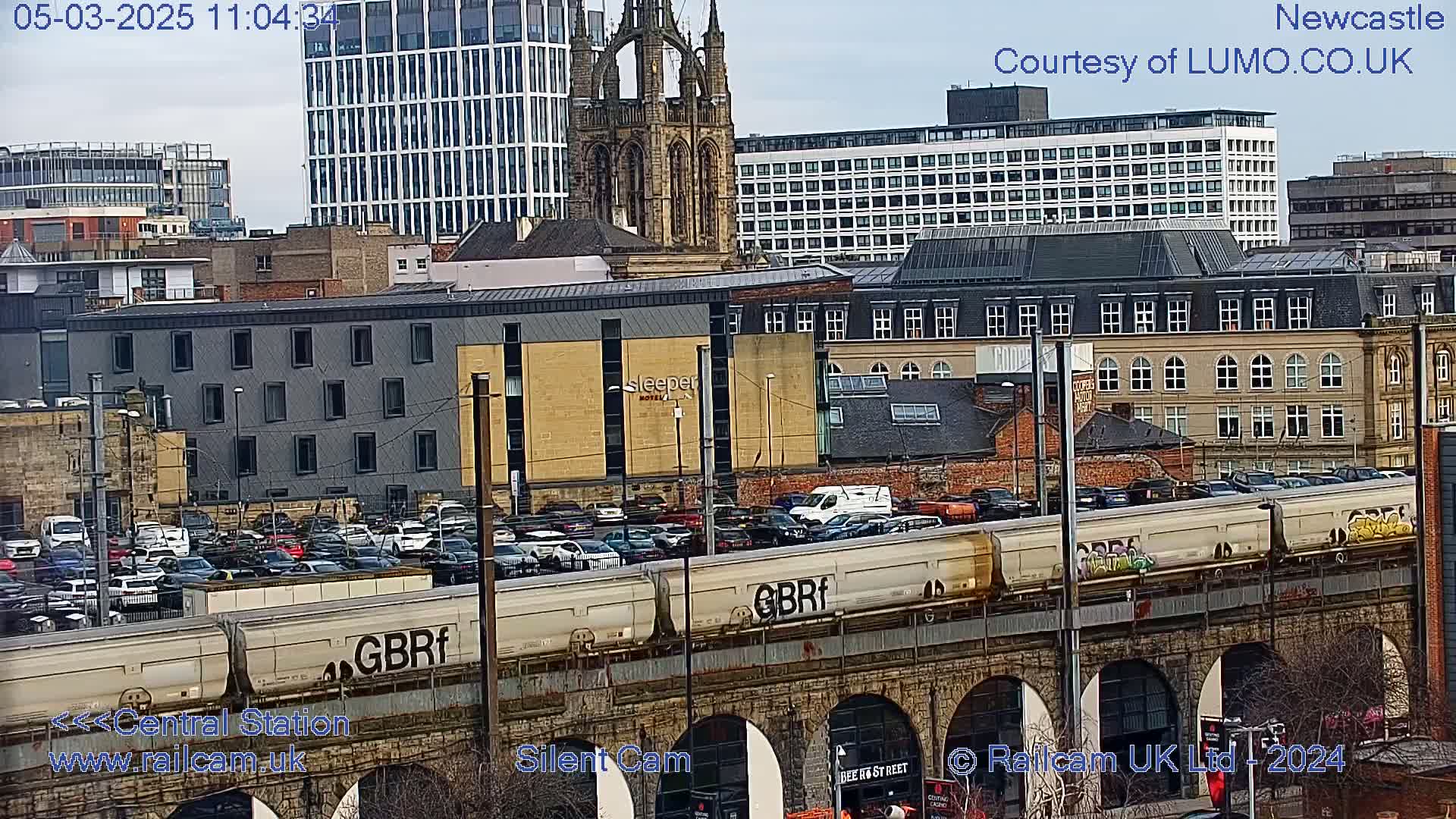 A freight train passes over a stone viaduct in a city, with overcast skies and numerous buildings, including a tall modern building and a Gothic-style church, visible in the background.