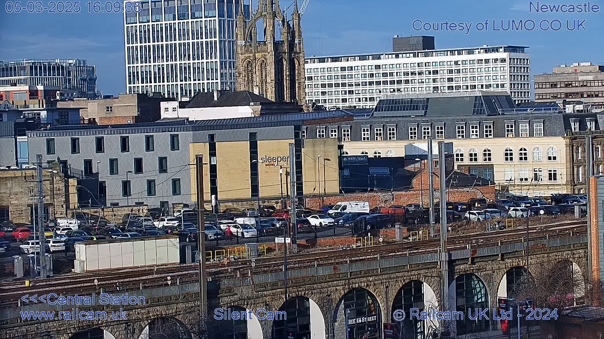 A sunny cityscape view shows a railway viaduct over a busy street with parked cars, various buildings including a hotel and a church, and a clear sky.