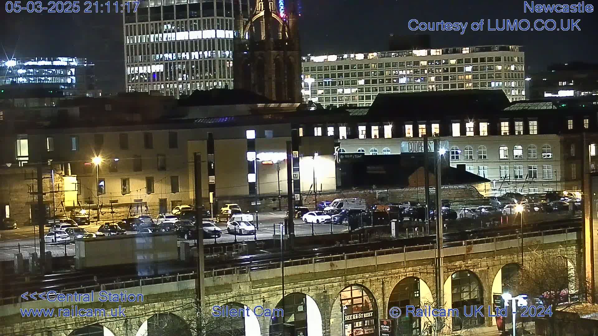 A nighttime view of a city’s central station, showing a parking lot full of cars, buildings with lit windows, and a stone railway viaduct.
