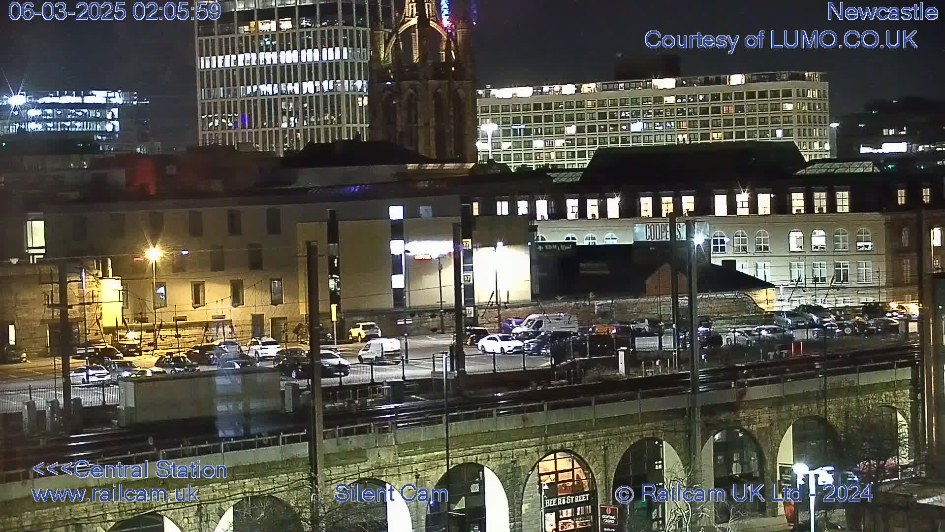 A nighttime view of a city, showing several buildings with illuminated windows, a parking lot with cars, and a train track over a stone archway.