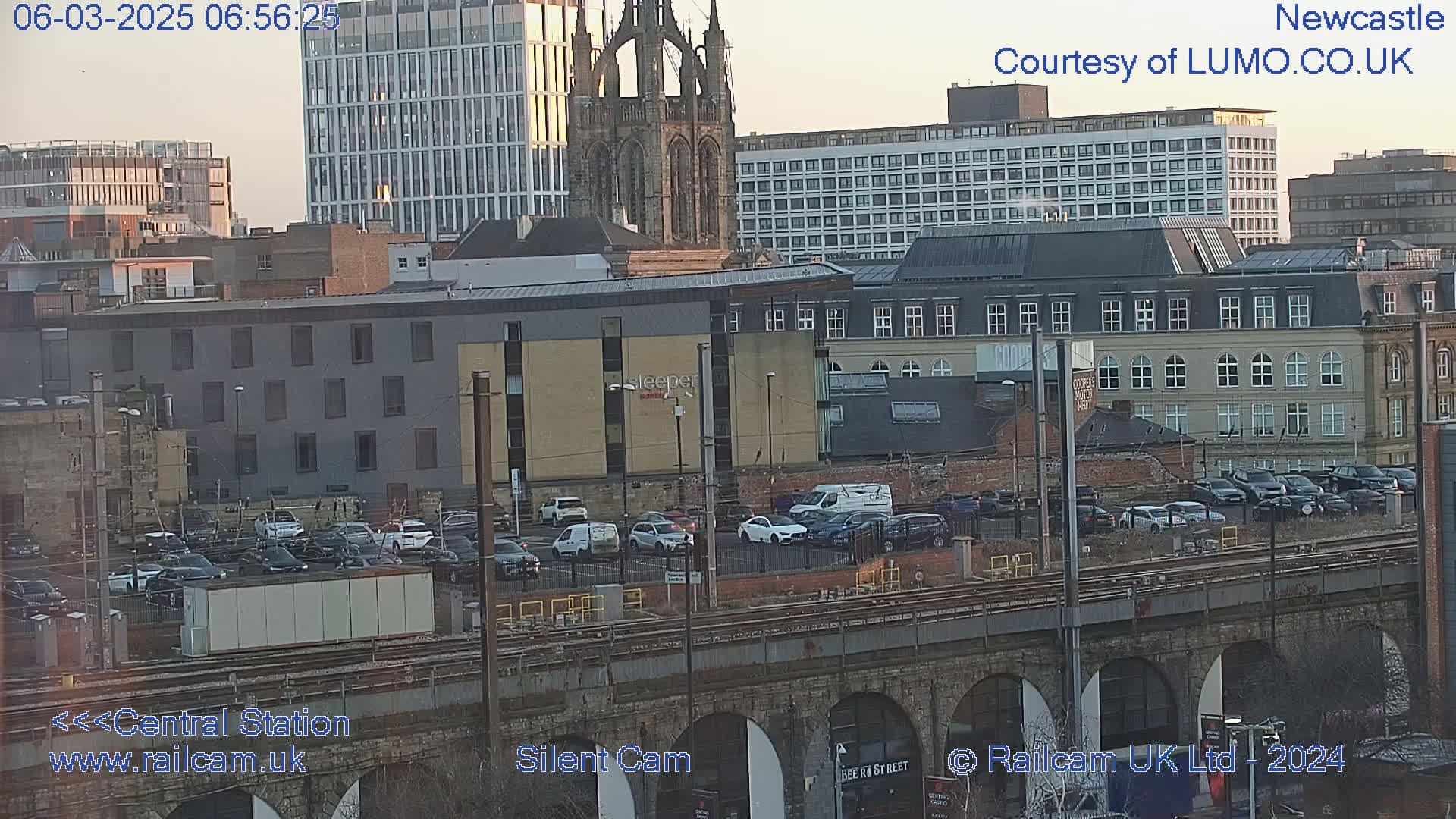 A city scene at sunset shows a railway bridge over a parking lot filled with cars, with modern and older buildings in the background.