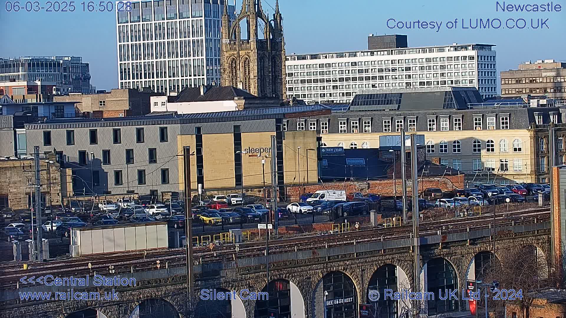 A sunny cityscape view shows a railway viaduct over a parking lot and various buildings, including a tall modern building and a cathedral, in the background.