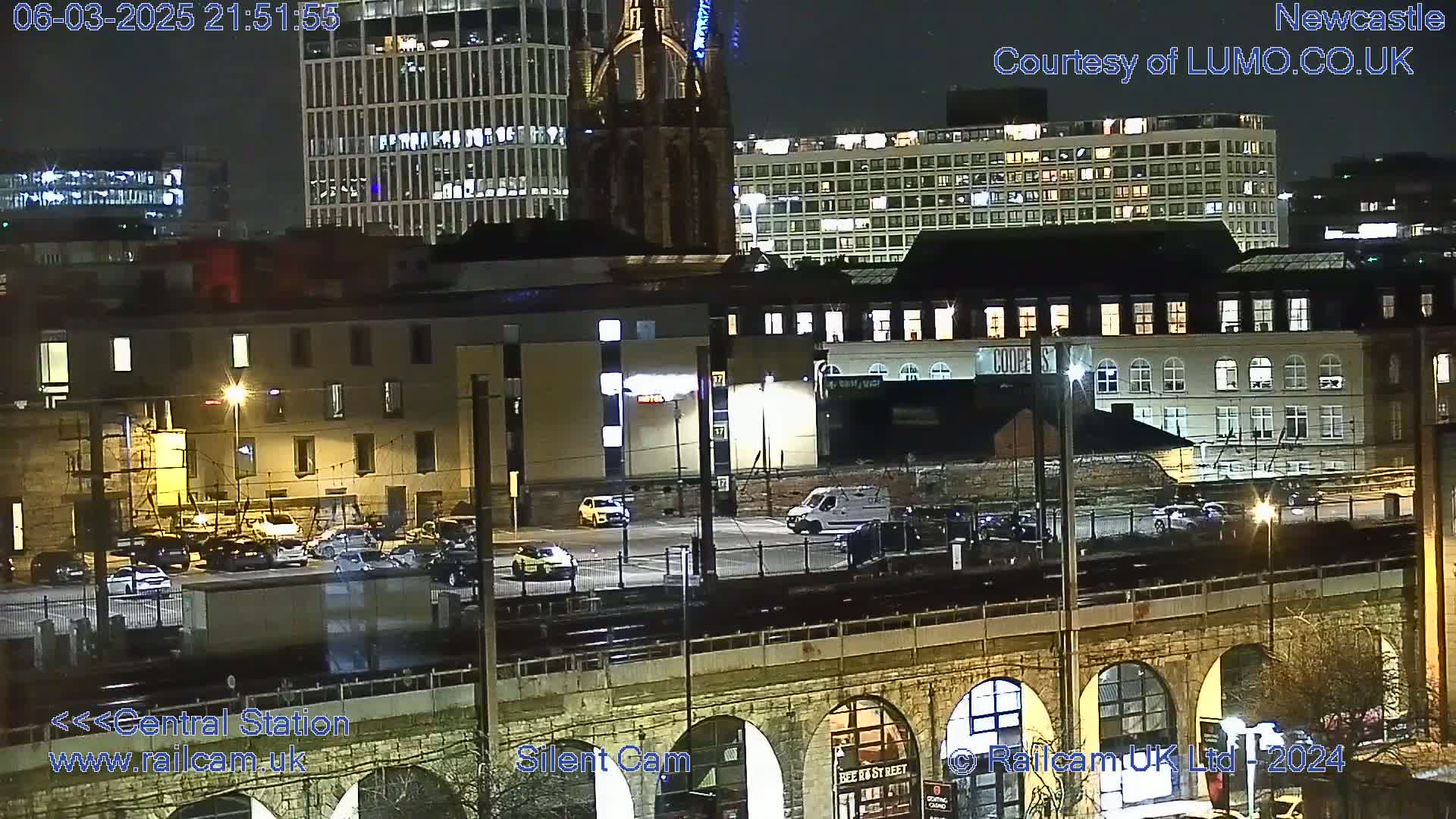 A nighttime view of a city with several buildings, a parking lot with cars, and a railway viaduct, under clear skies.