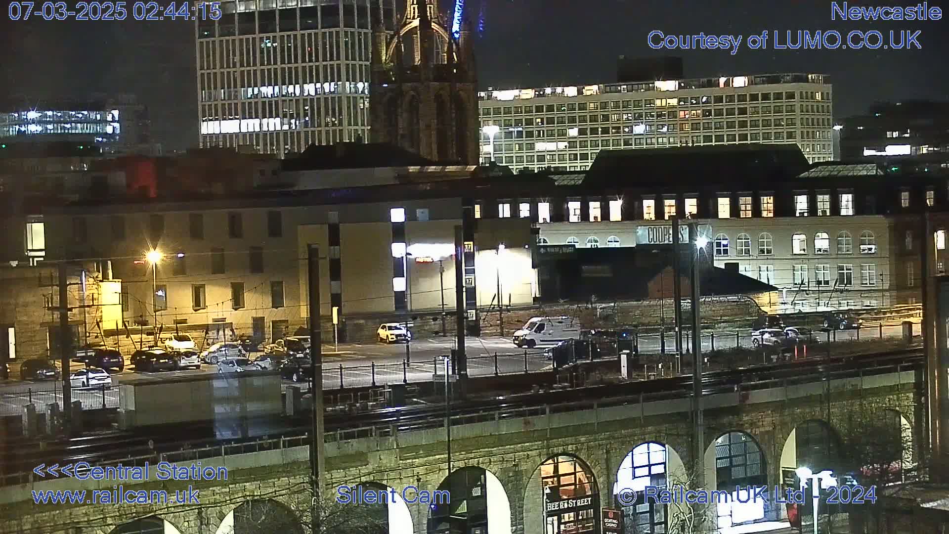 A nighttime view of a city, showing several buildings, a parking lot with cars, train tracks, and a stone archway bridge, under clear skies.