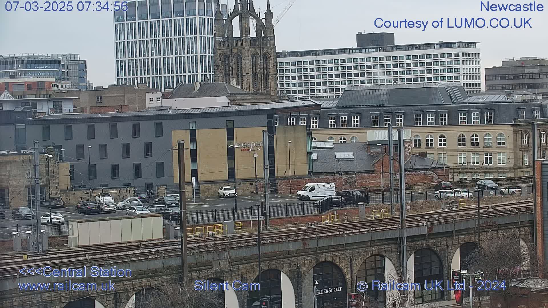 A view of a city's buildings and a parking lot on an overcast day, with train tracks and a stone bridge visible in the foreground.