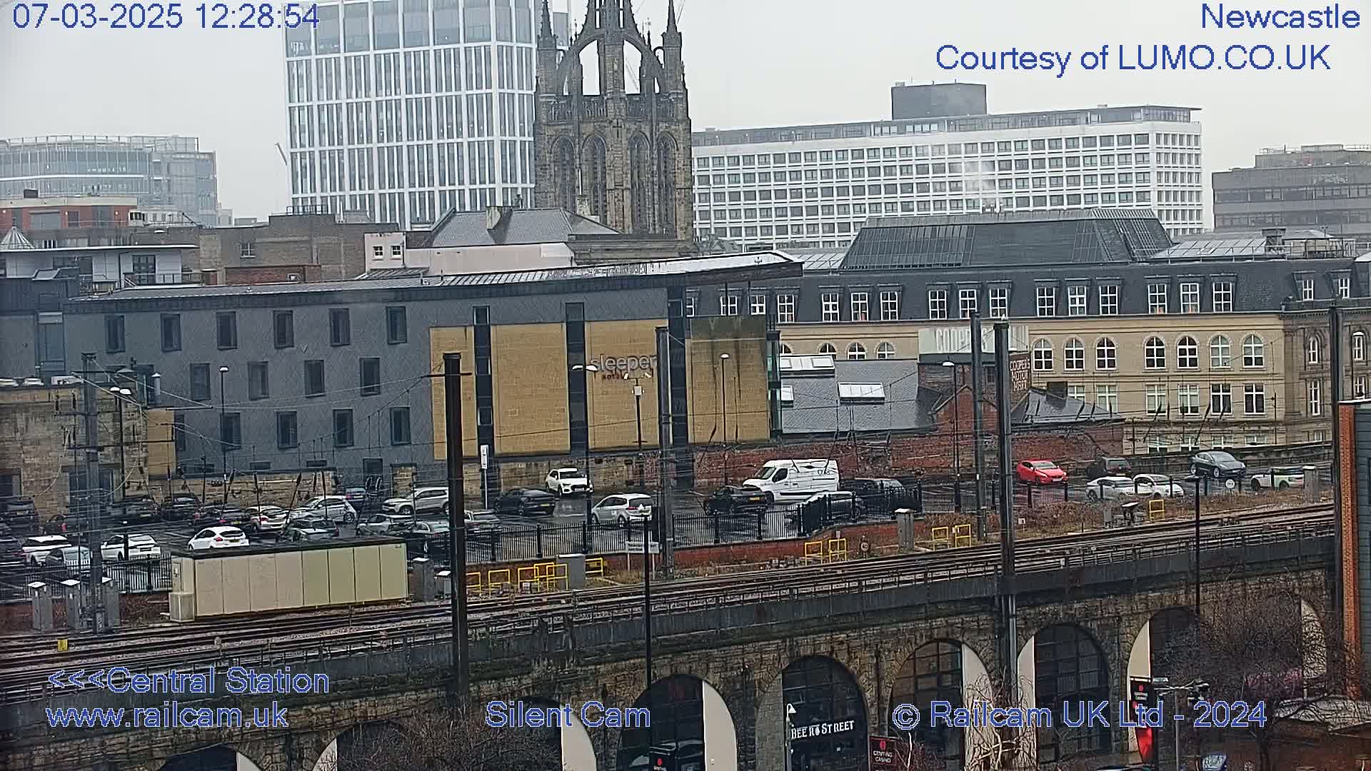 A high-angle, long shot shows a city streetscape on an overcast day, with railway tracks running through the foreground and various buildings, including a large church, in the background.