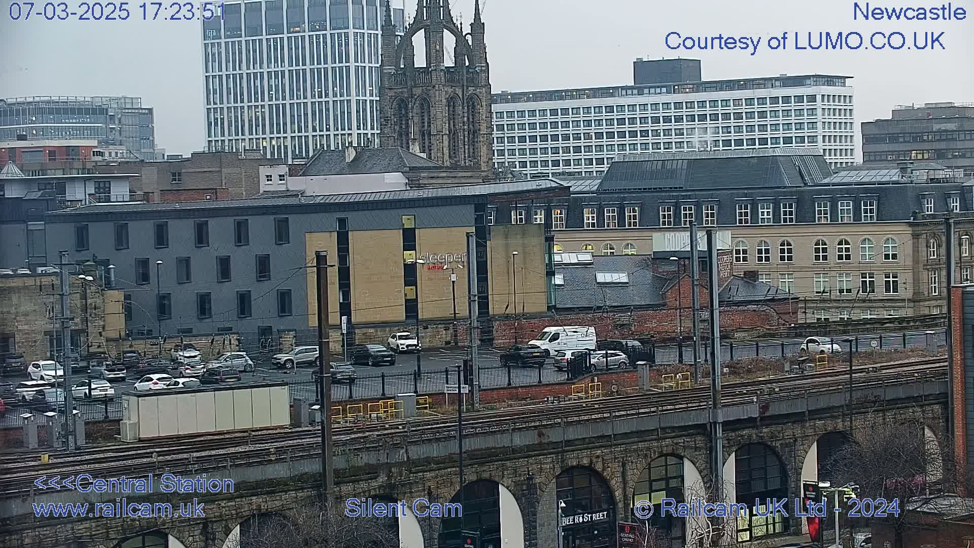 A city scene viewed from above, showing a mix of modern and older buildings, a parking lot with cars, train tracks, and a stone viaduct, under an overcast sky.