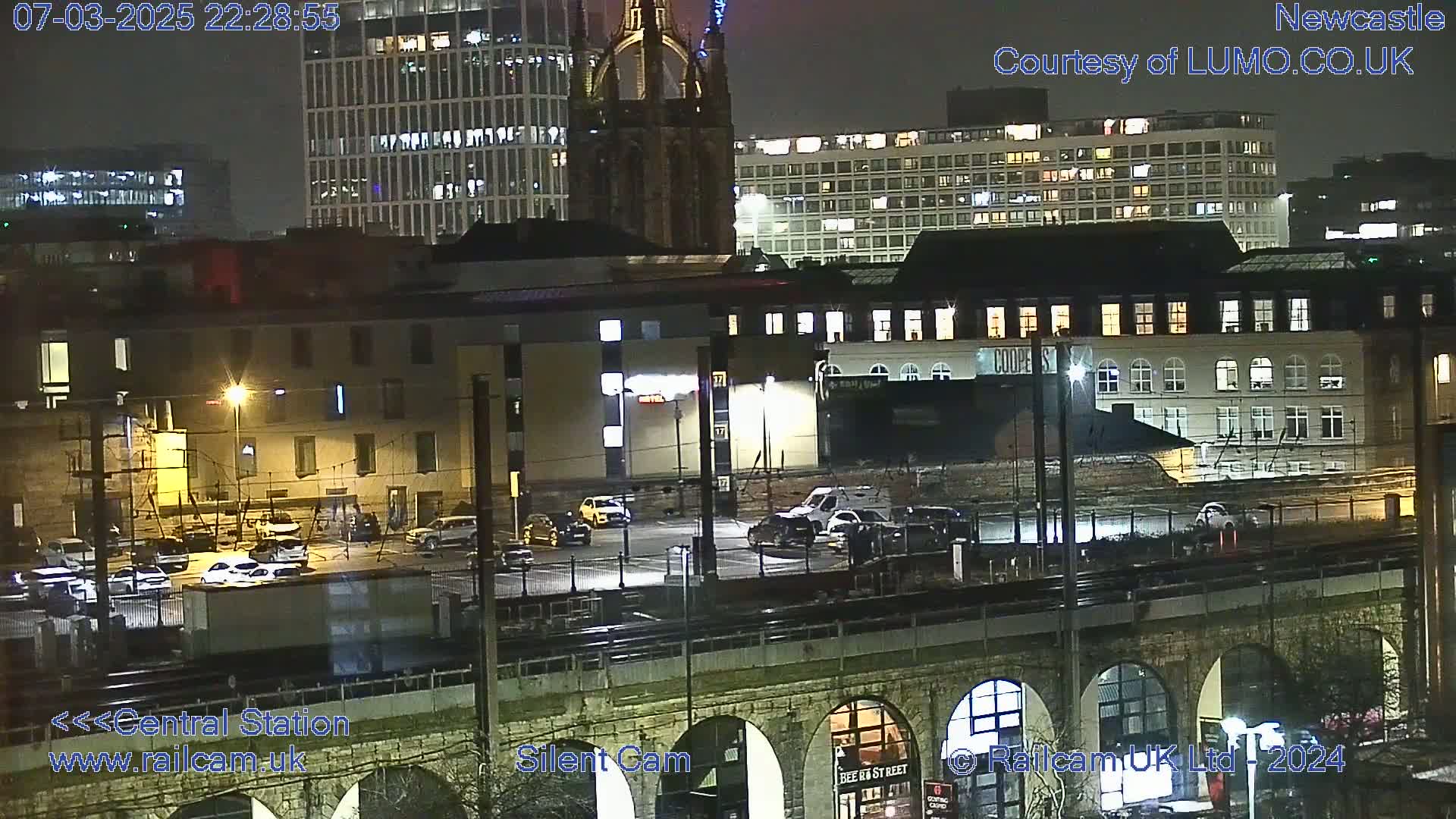 A nighttime view of a city's cityscape, including buildings, cars parked in a lot, and a train trestle, under clear skies.