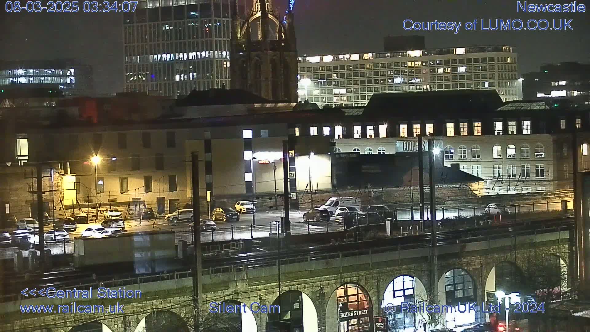 A nighttime view of a city, showing buildings, a parking lot with cars, and a railway viaduct under a clear sky.