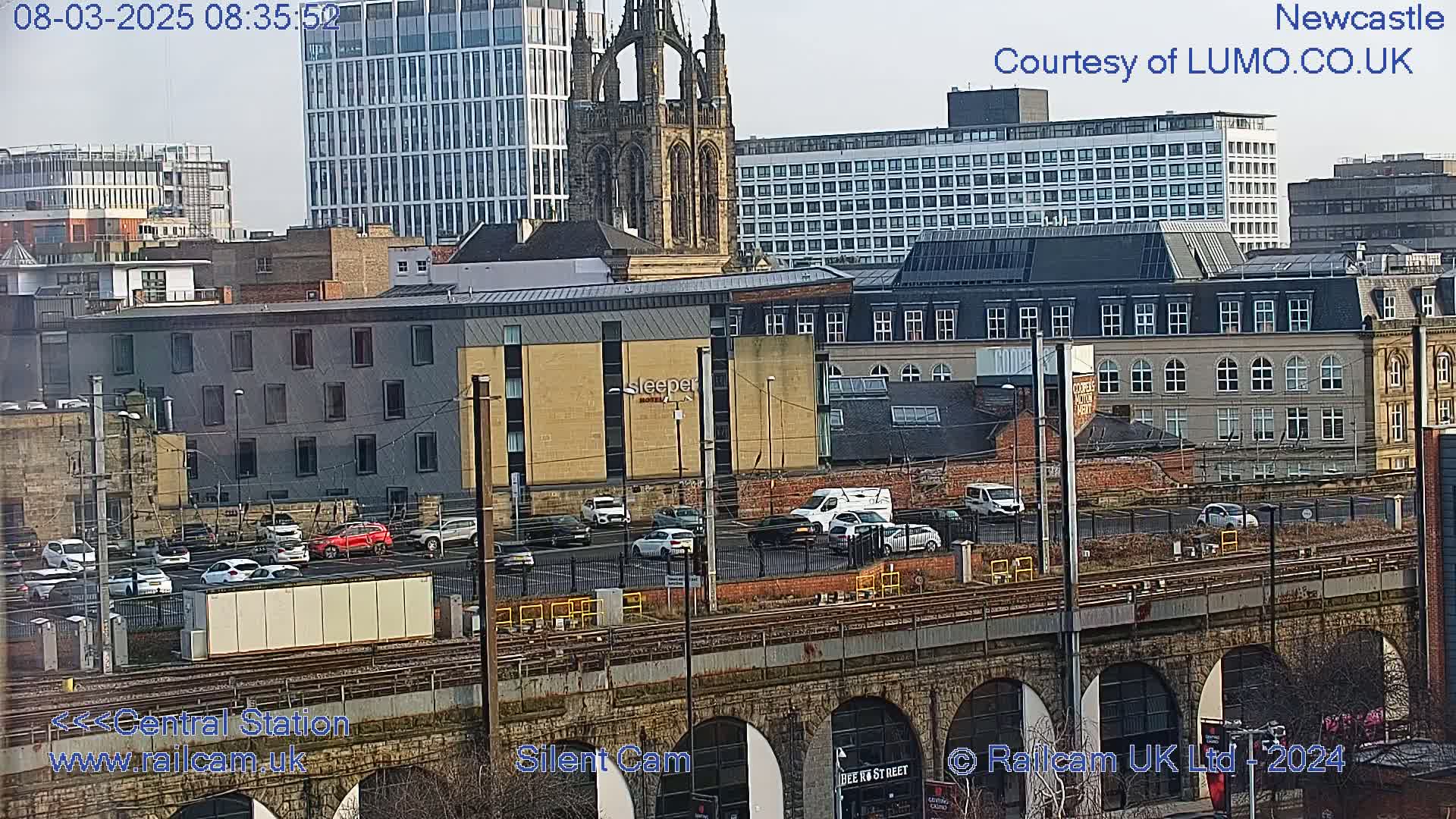 A city view on a cloudy day shows several buildings, including a tall modern building and a church, with cars parked in a lot near train tracks and a stone underpass.