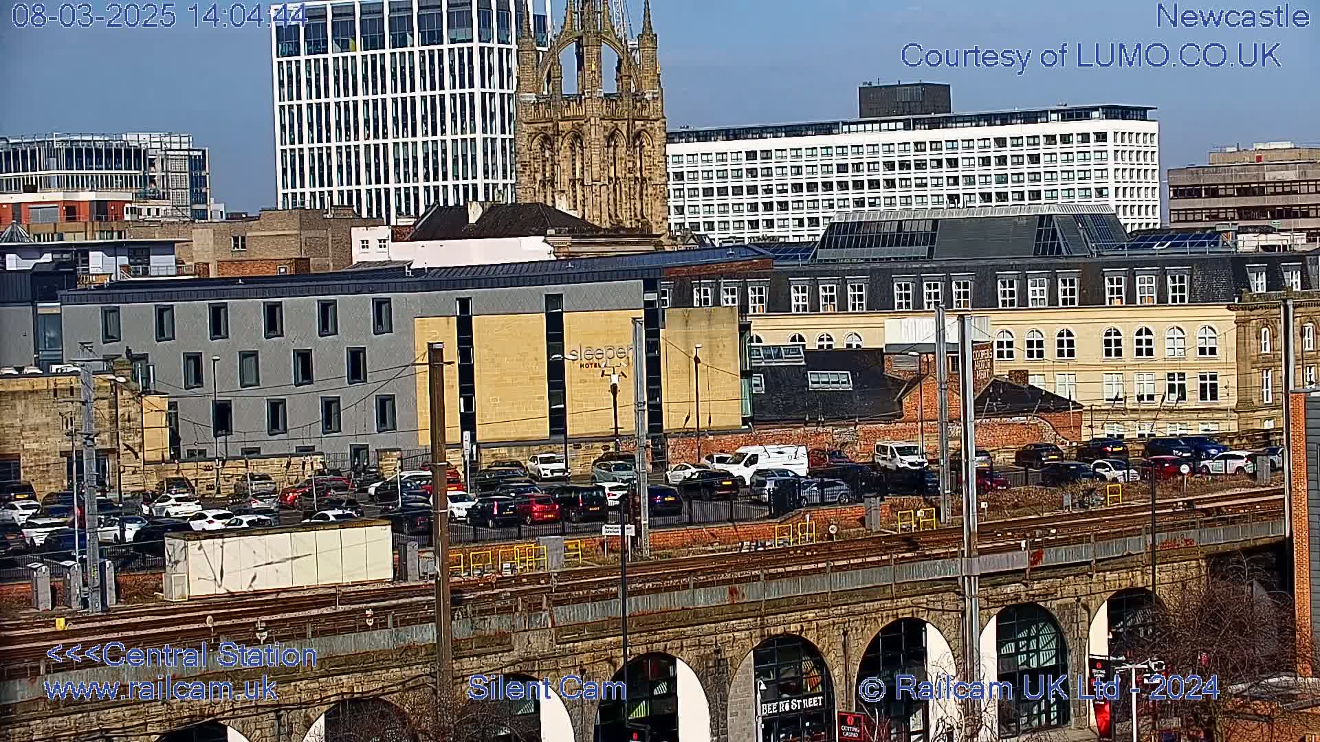 A city scene on a sunny day, viewed from above a railway viaduct, shows numerous buildings including a large church, a hotel, and apartment blocks, with cars parked in a lot and traffic on a road below.