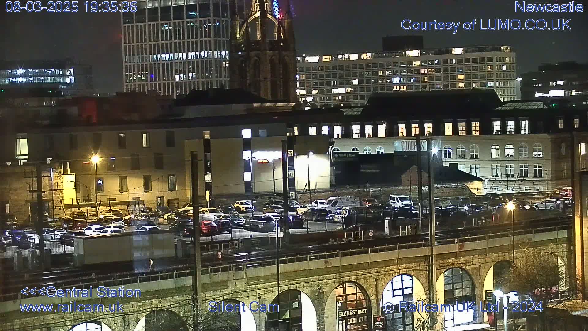 A nighttime view of a city's central station area, showing parked cars, buildings with illuminated windows, and a stone bridge with arches.