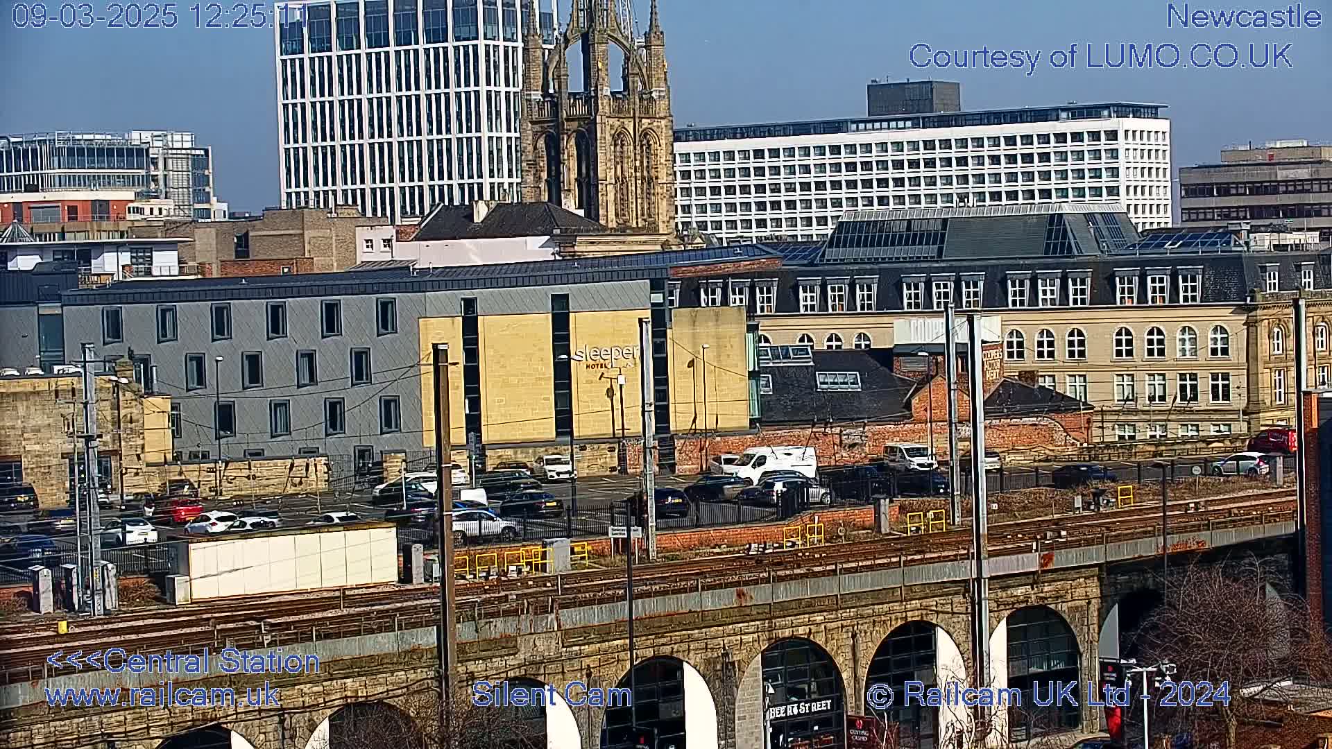A sunny cityscape view shows a railway viaduct over a street with parked cars and buildings, including a church spire and modern high-rises.