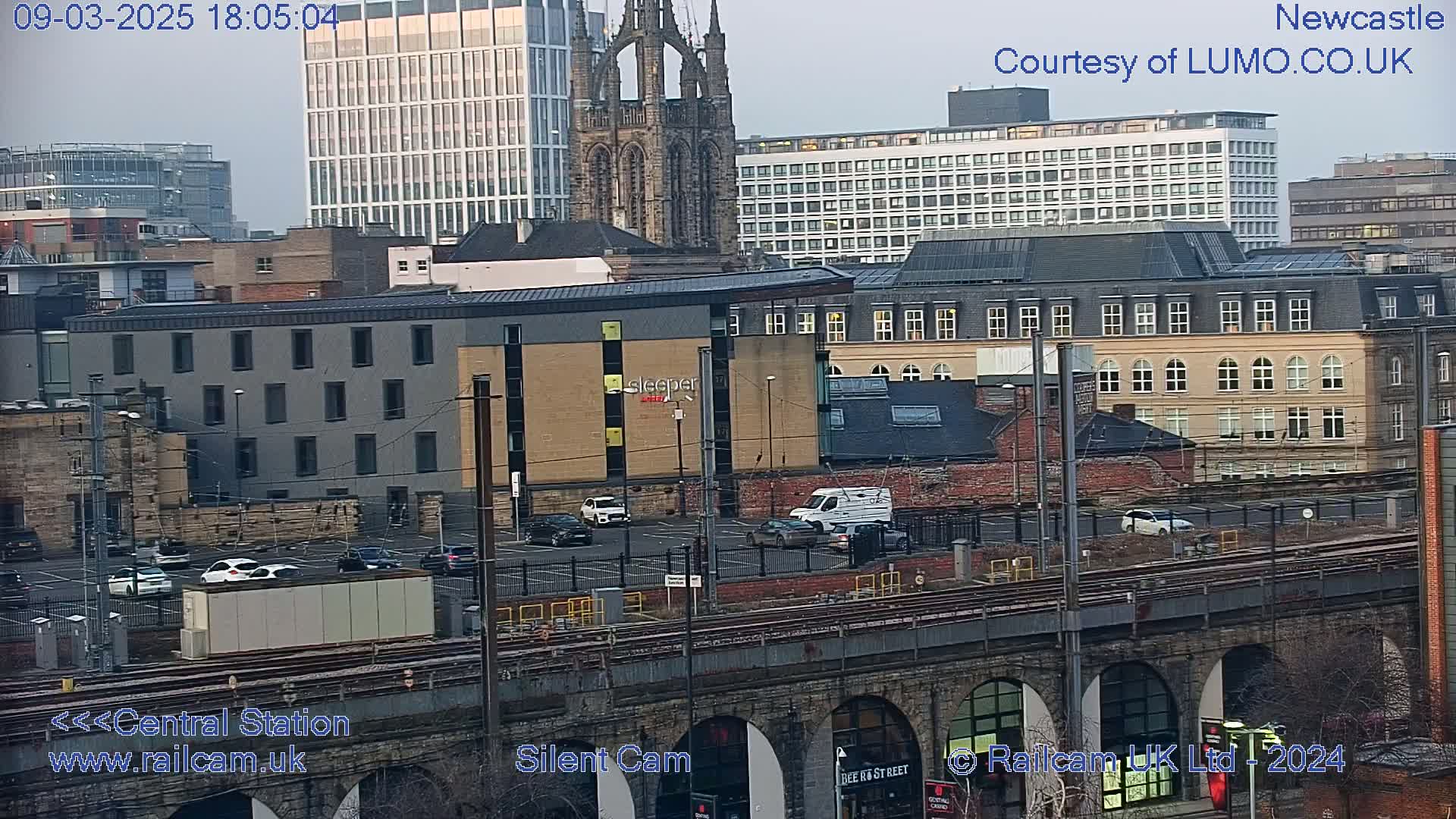 A city landscape at dusk showing a mix of modern and older buildings, a parking lot with cars, train tracks, and a stone bridge.