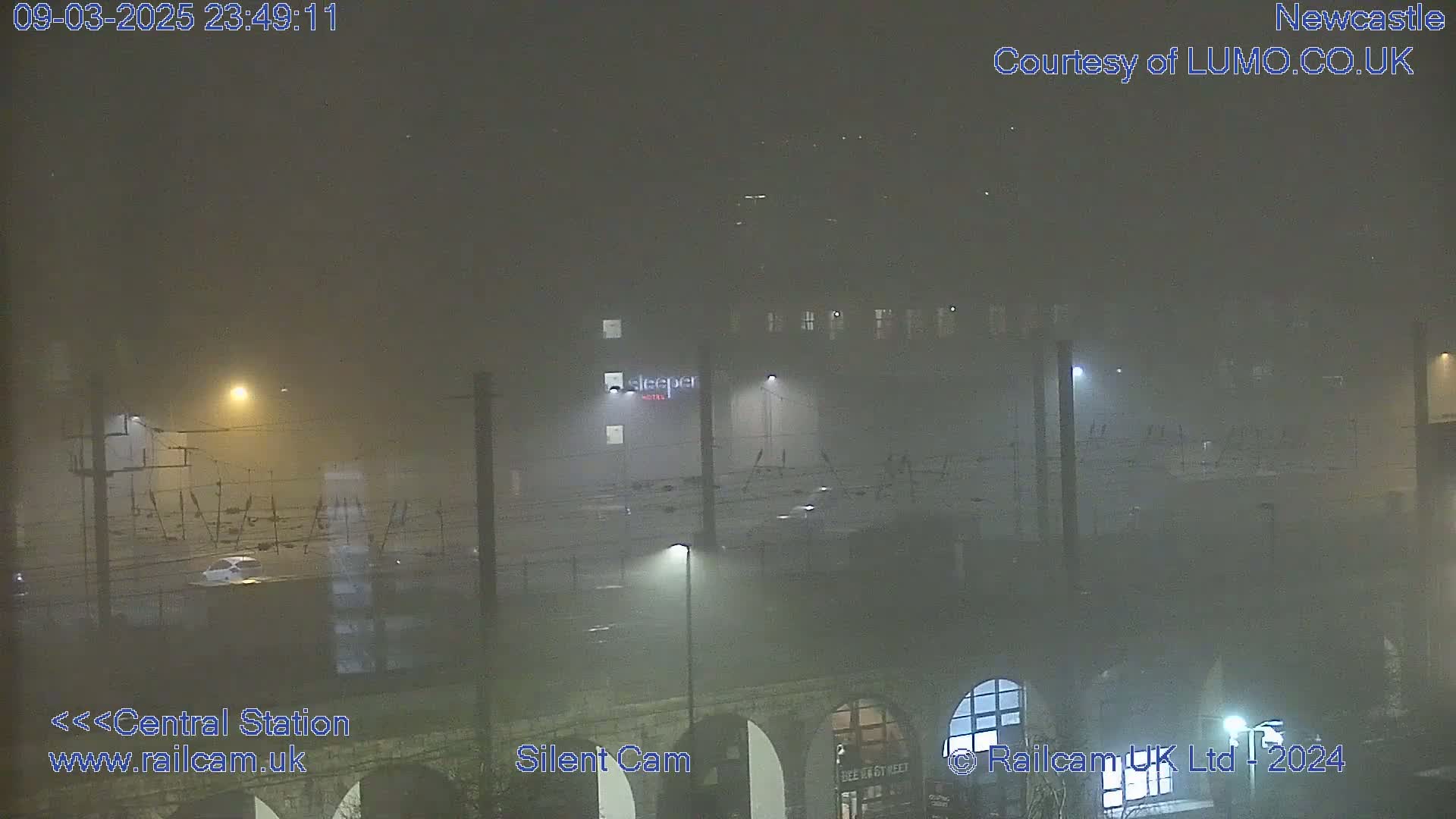 A nighttime, foggy view of a city area showing a parking lot, a building with lights, and overhead power lines.