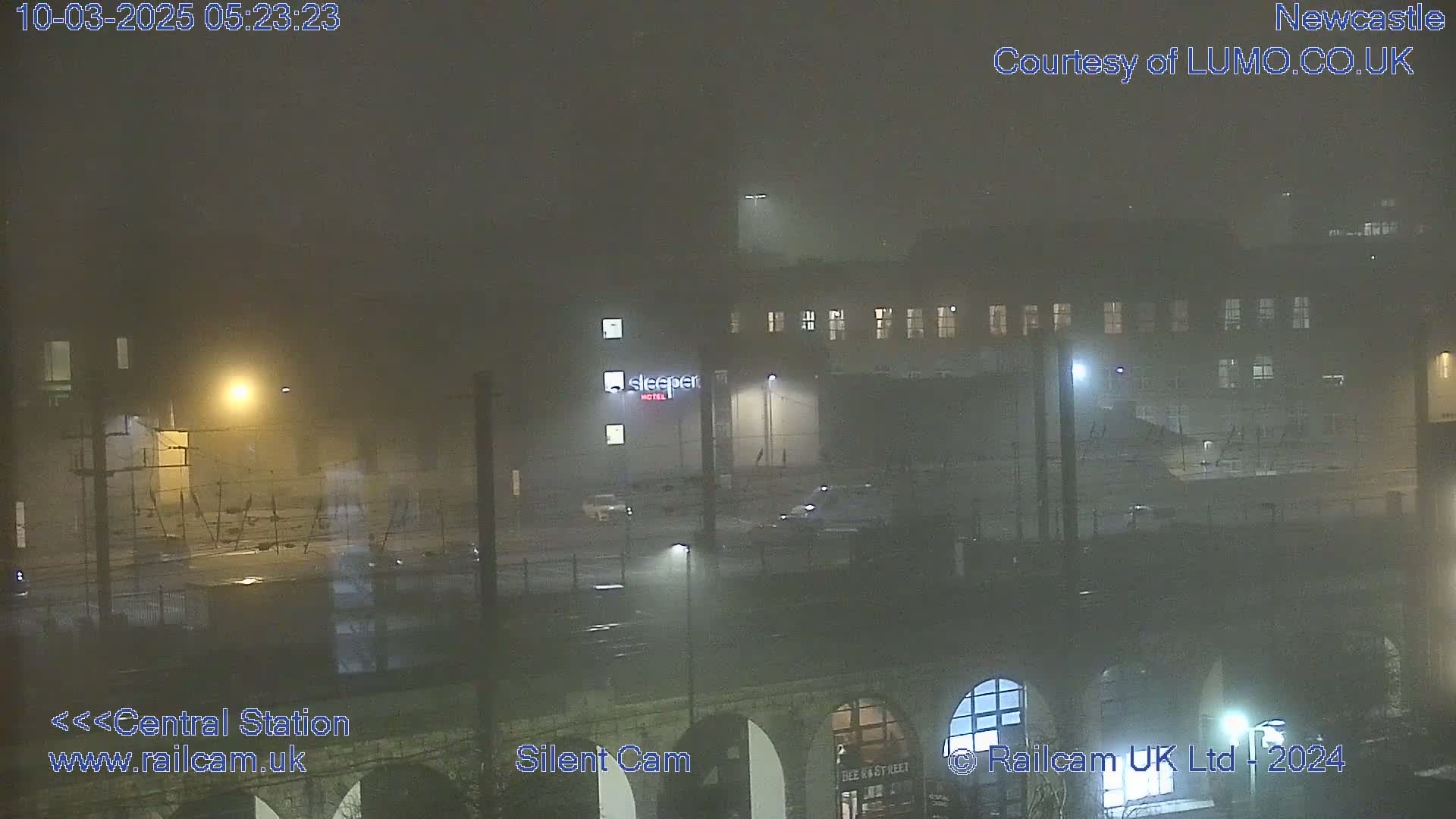A foggy night scene shows a multi-story building with lights on, a street, and a bridge.