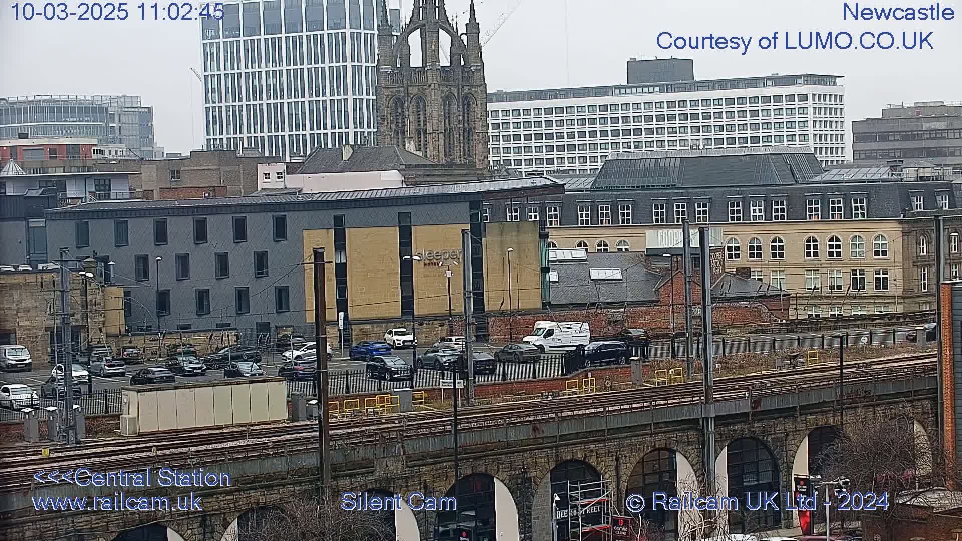 A city scene on an overcast day, viewed from a high vantage point, shows a railway viaduct, a parking lot filled with cars, and numerous buildings of varying architectural styles, including a tall modern structure and an older church-like building.