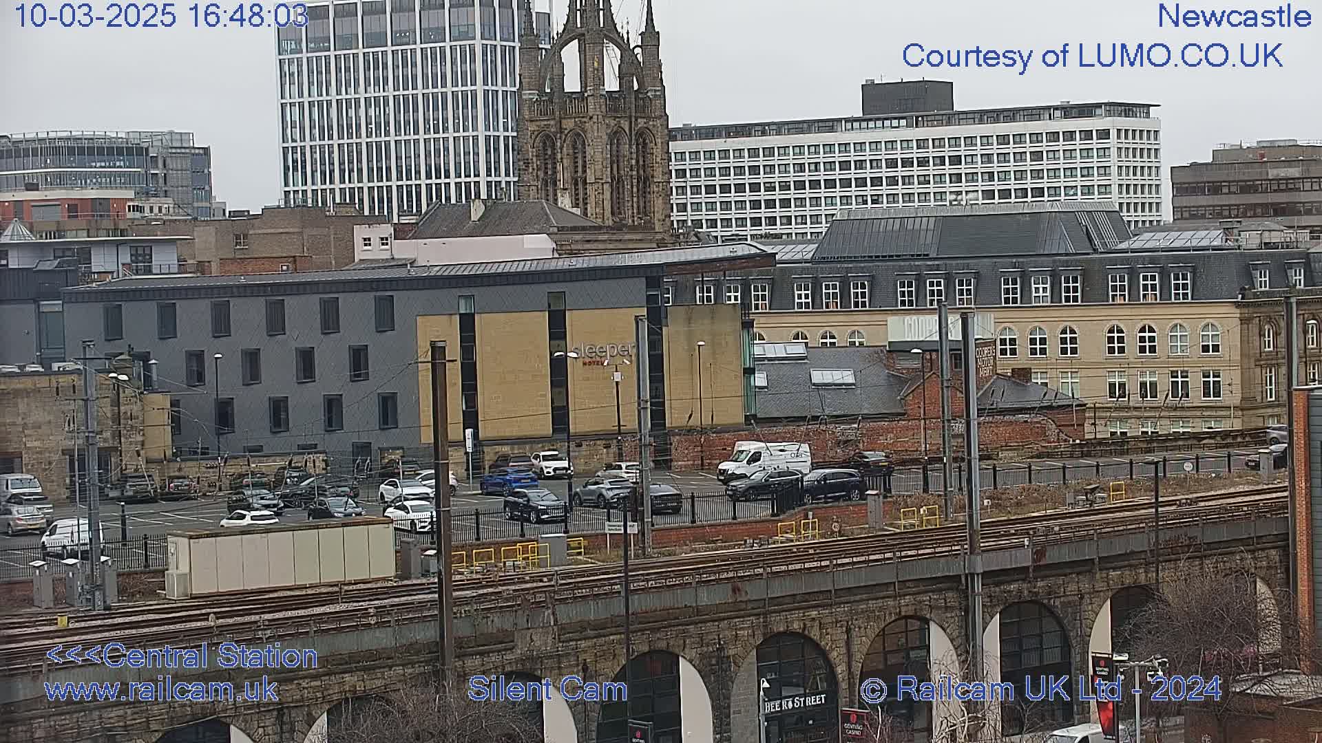 A city view on an overcast day, showing a parking lot, train tracks on a stone viaduct, and various buildings including a tall modern building and an older church.