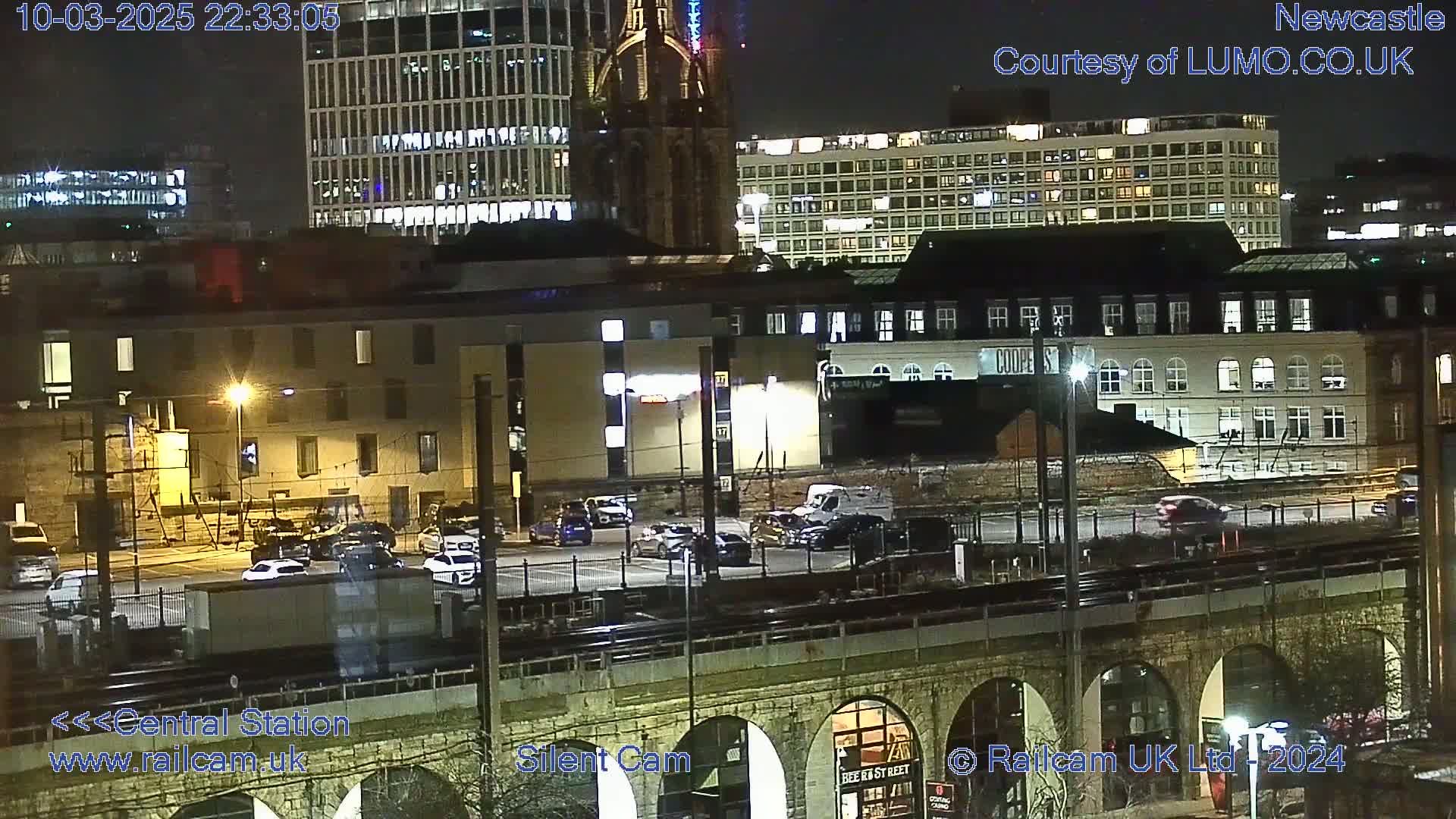 A nighttime view of a city featuring several buildings, a parking lot with cars, and a railway viaduct.