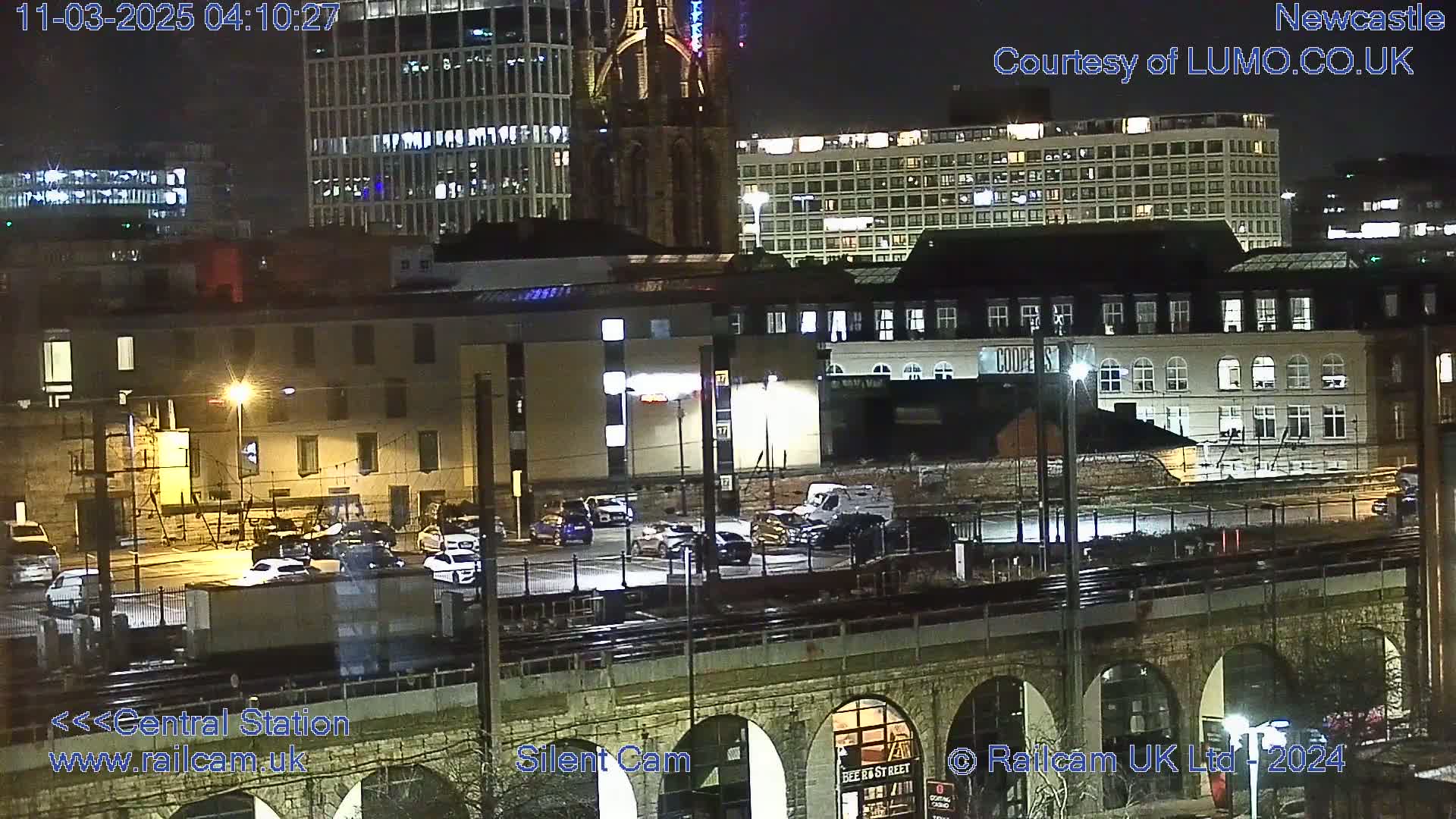 A nighttime view of a city's cityscape, featuring buildings, vehicles in a parking lot, and train tracks spanning an arched bridge.