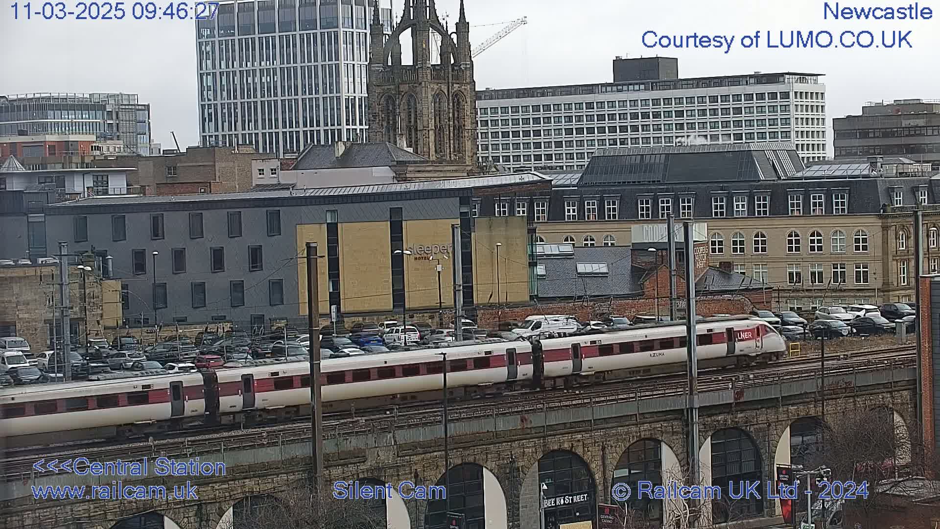 A long white and maroon passenger train travels across a stone bridge over a parking lot and city buildings under an overcast sky.