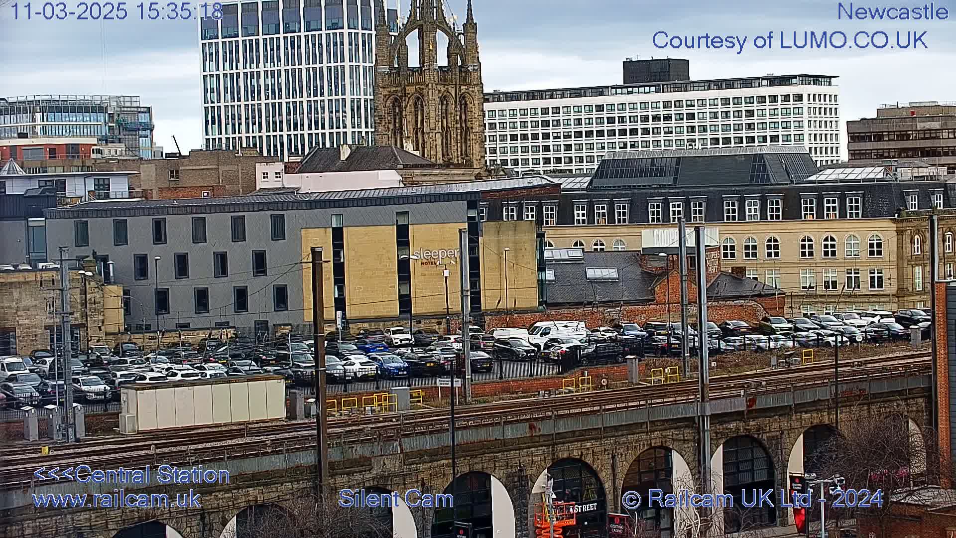 A city view on an overcast day shows a large parking lot next to a railway viaduct, with various buildings including a church and high-rises in the background.