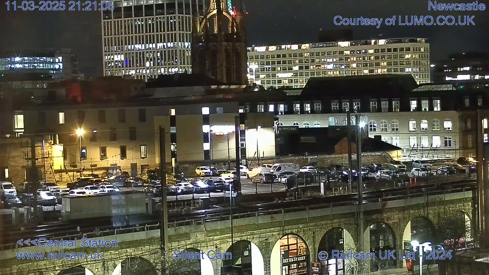 A nighttime view of a city, showing several buildings, a parking lot full of cars, and a train track elevated on a stone structure.