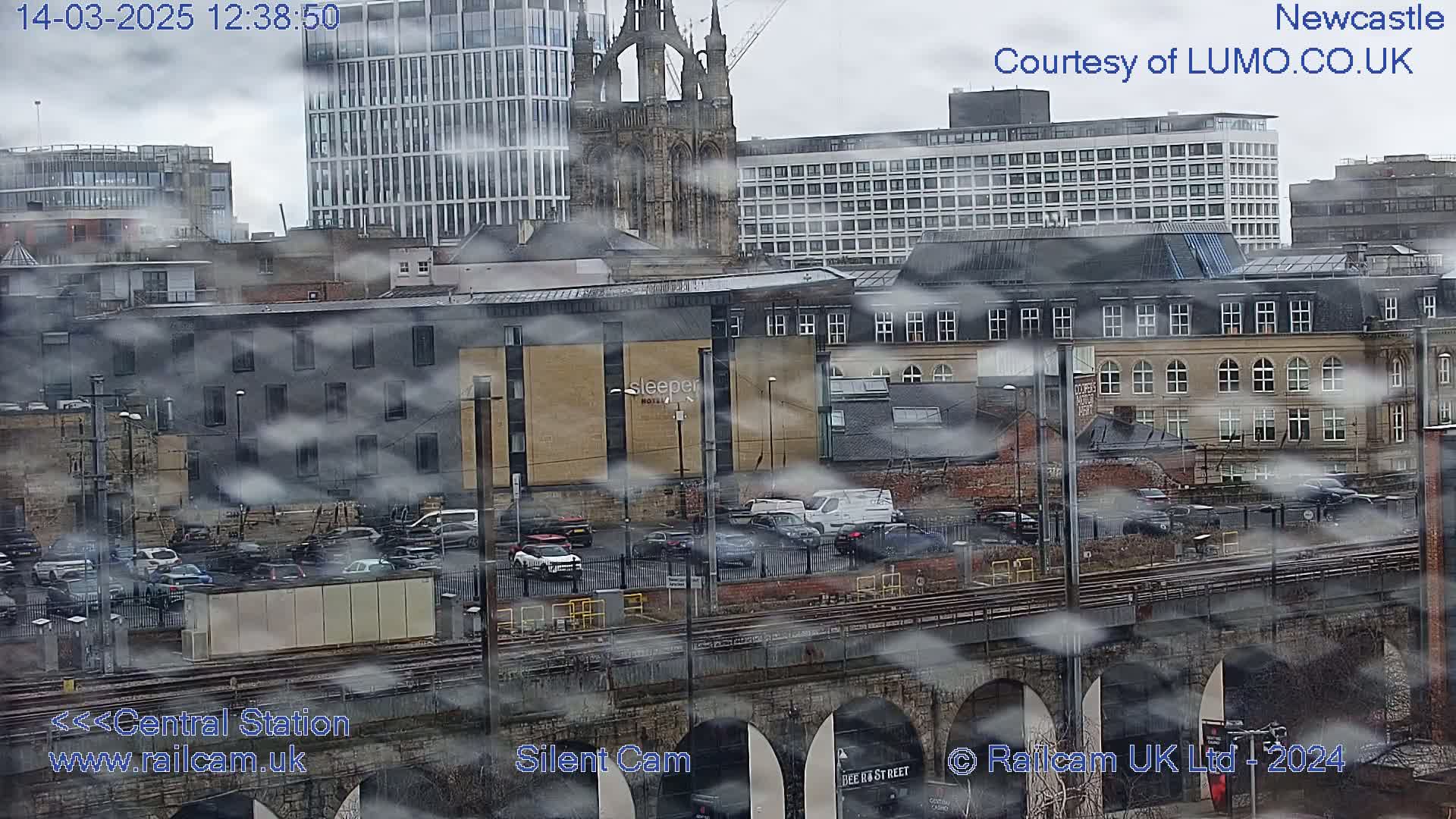 A city scene viewed from above on a cloudy day, showing buildings, vehicles, and train tracks, with a low-lying fog or mist obscuring parts of the view.