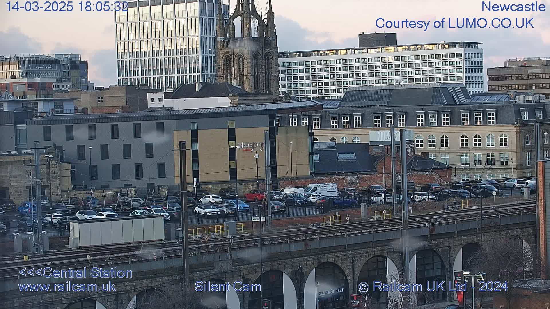A city skyline with overcast skies, featuring numerous buildings, a train station, and a parking lot filled with cars, all viewed from an elevated vantage point.