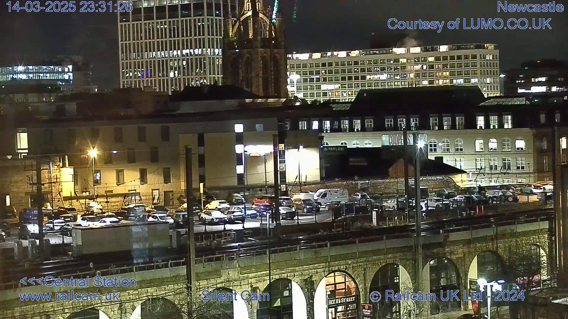 A nighttime view of a city's illuminated buildings and a parking lot near a railway overpass, under a clear sky.