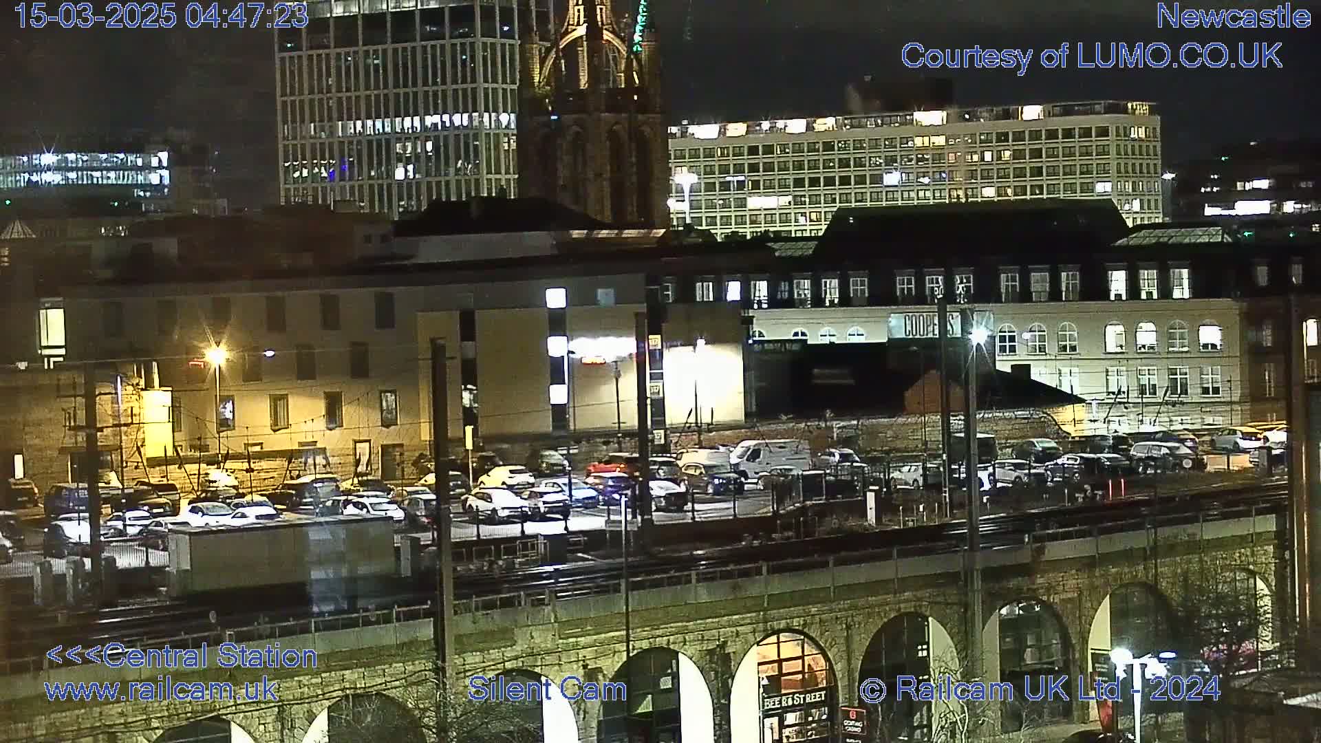 A nighttime view of a city's central station area, showing a parking lot full of cars, buildings with illuminated windows, and a stone bridge archway.
