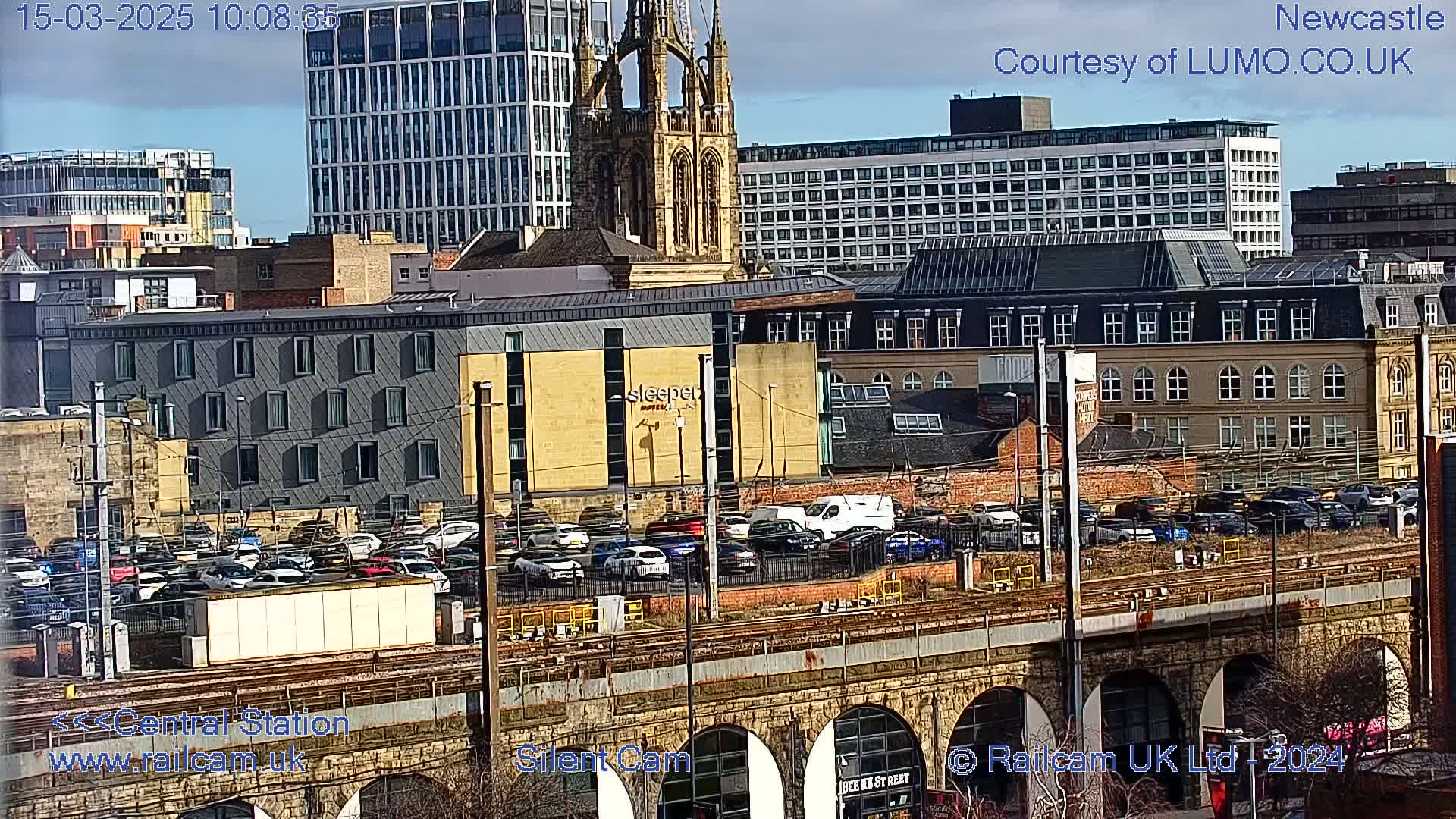 A city scene on a partly sunny day, viewed from above a train trestle, shows a mix of modern and older buildings, including a large church, a parking lot full of cars, and a hotel.