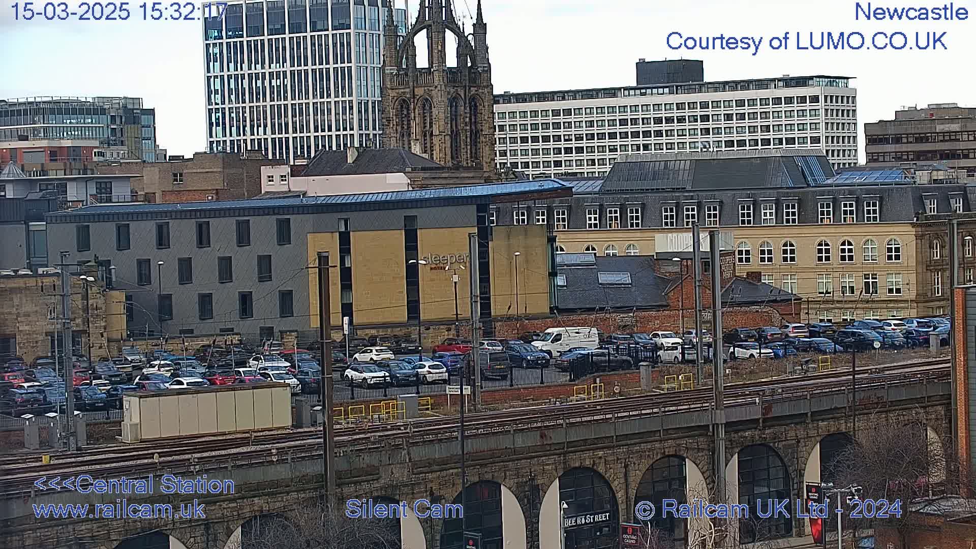 A large parking lot filled with cars sits below a railway bridge and overlooks a cityscape of various buildings including a tall modern building, a church, and older brick structures, all under an overcast sky.